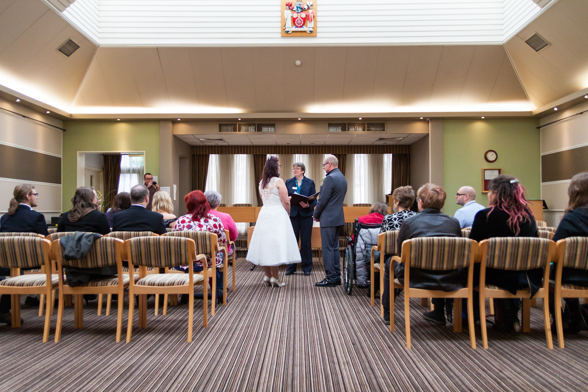 A wedding ceremony. Taken from the back of the room. The bride and groom stand at the front of the aisle facing the registrar.