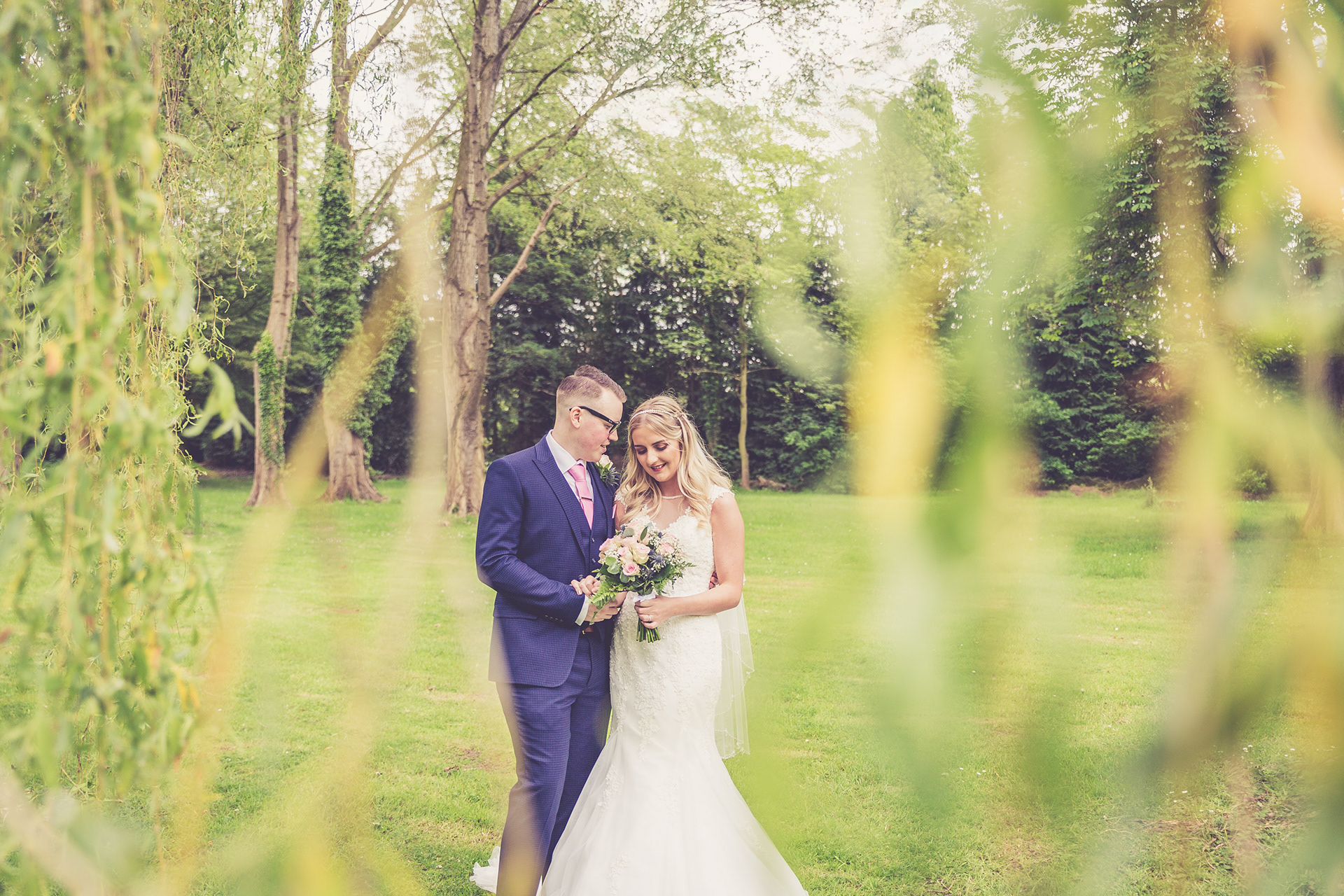 North Wales Wedding Photographer. Portrait of a bride and groom on their wedding day. They are stood in the lovely gardens of MacDonald Craxton Wood Hotel and Spa. The groom is wearing a dark blue suit and pink tie. The bride has a long fitted wedding dress which flares from around her knees.