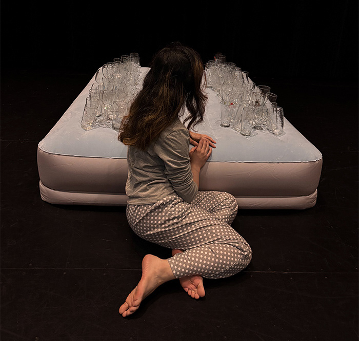 A performer kneels beside a mattress covered with glass tumblers in Deep Sleep, a multidisciplinary performance and installation by Armin Shohrati.