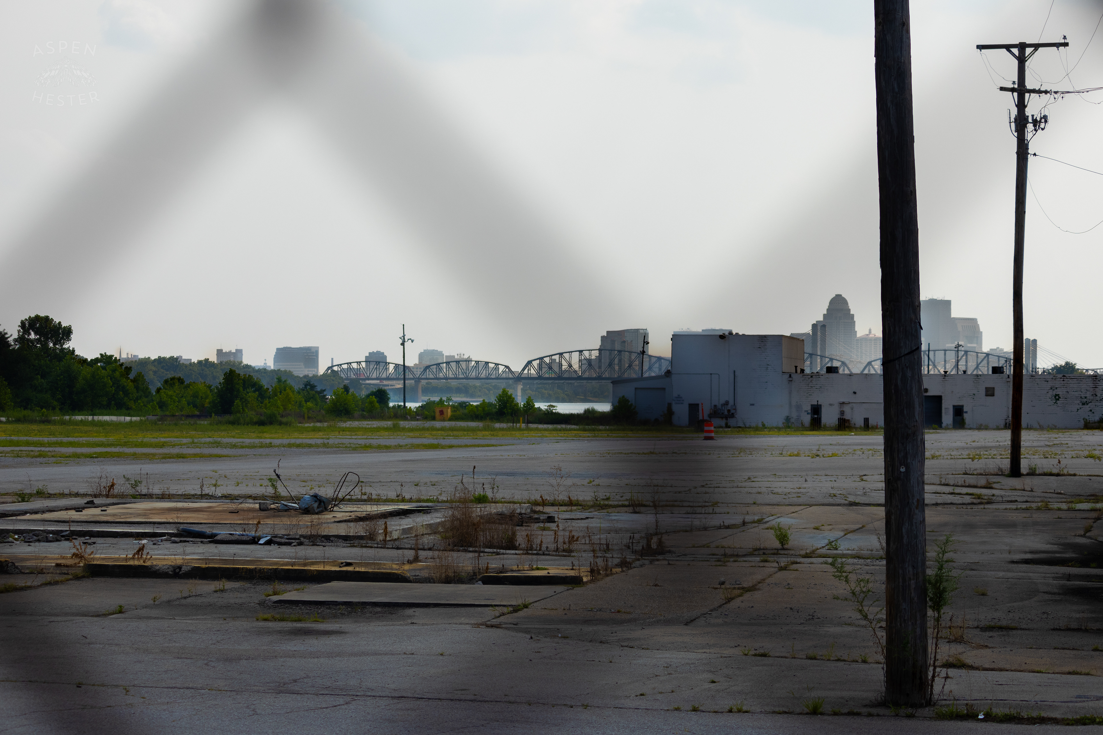 Looking Through the Chain Link Fence Towards the Louisville Skyline at the Abandoned Jeffboat Shipyard. July 26th, 2024/Aspen Hester