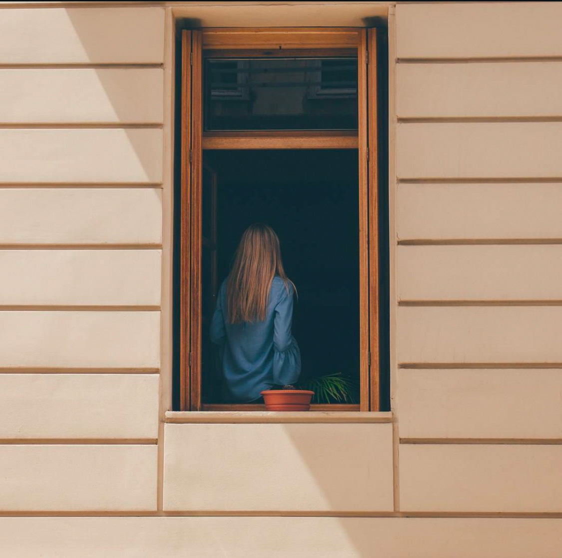 A solitary figure standing at a window, illuminated by natural light, framed by architectural simplicity.