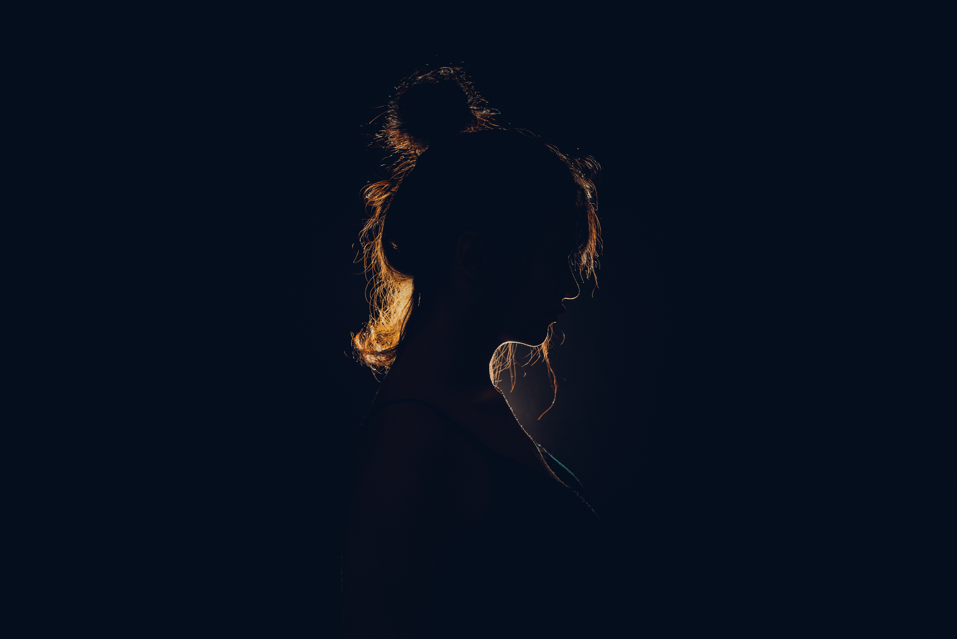 Silhouette of a woman with her hair tied in a bun, highlighted by a soft, warm backlight that outlines her profile against a dark background, creating a dramatic and contemplative mood.