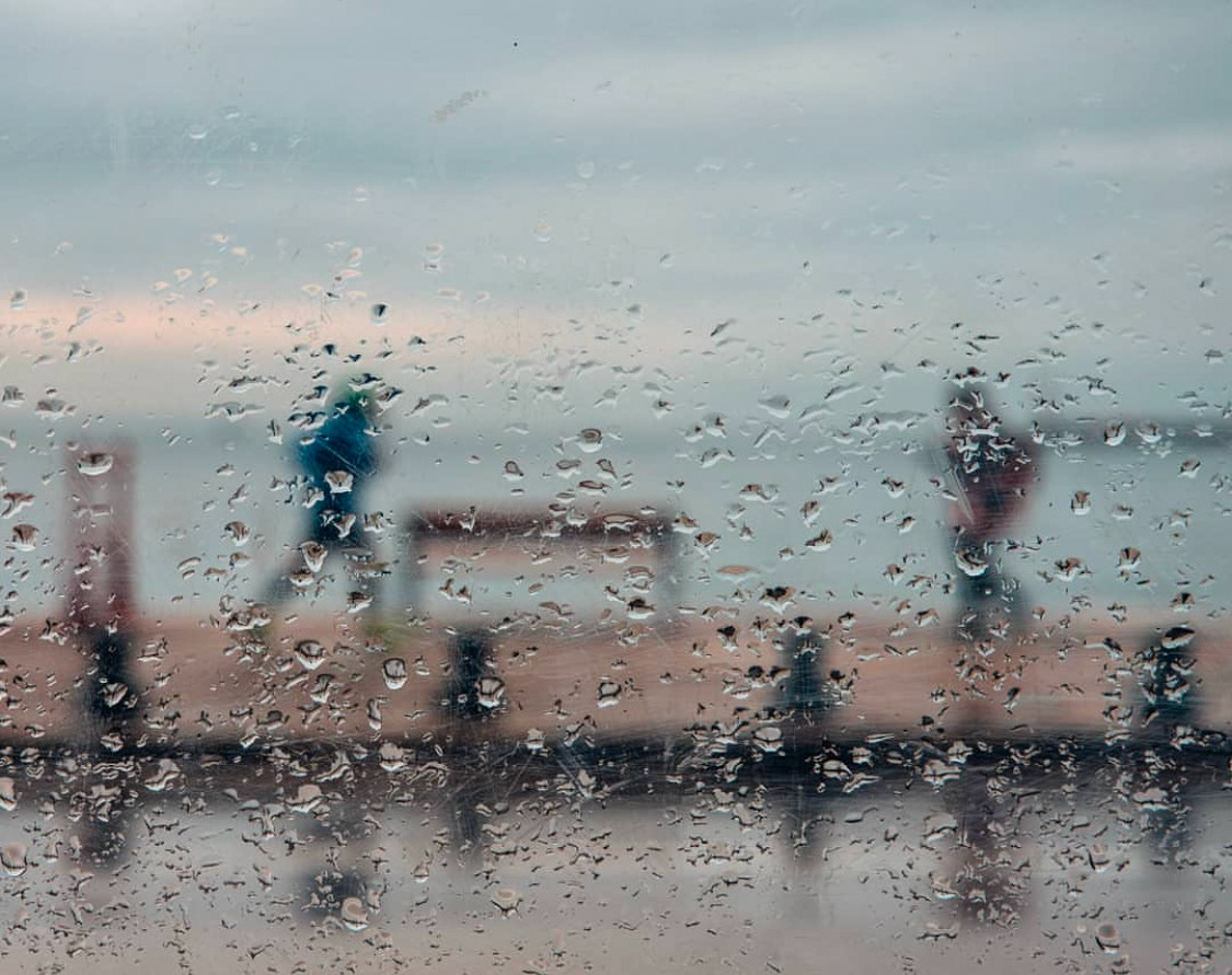 A rainy window with visible droplets blurs the silhouettes of people walking by the waterfront in the background.