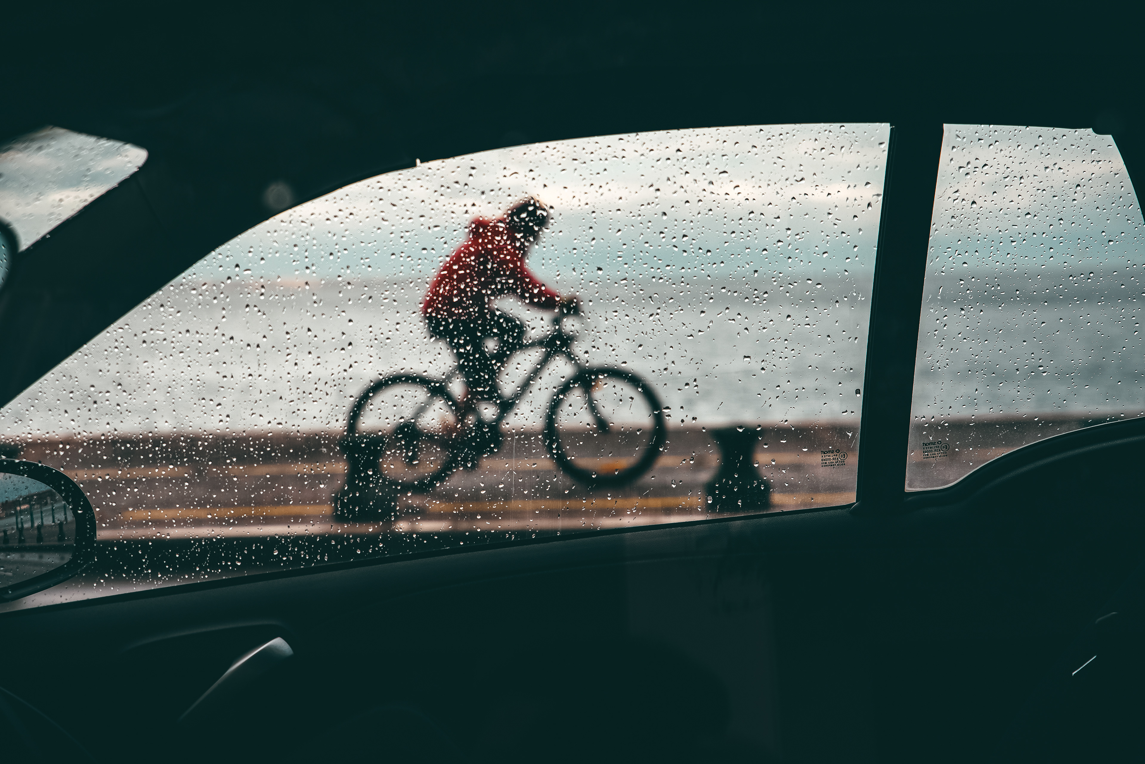 A cyclist in a red jacket seen through a rain-speckled car window, with blurred motion enhancing the sense of movement