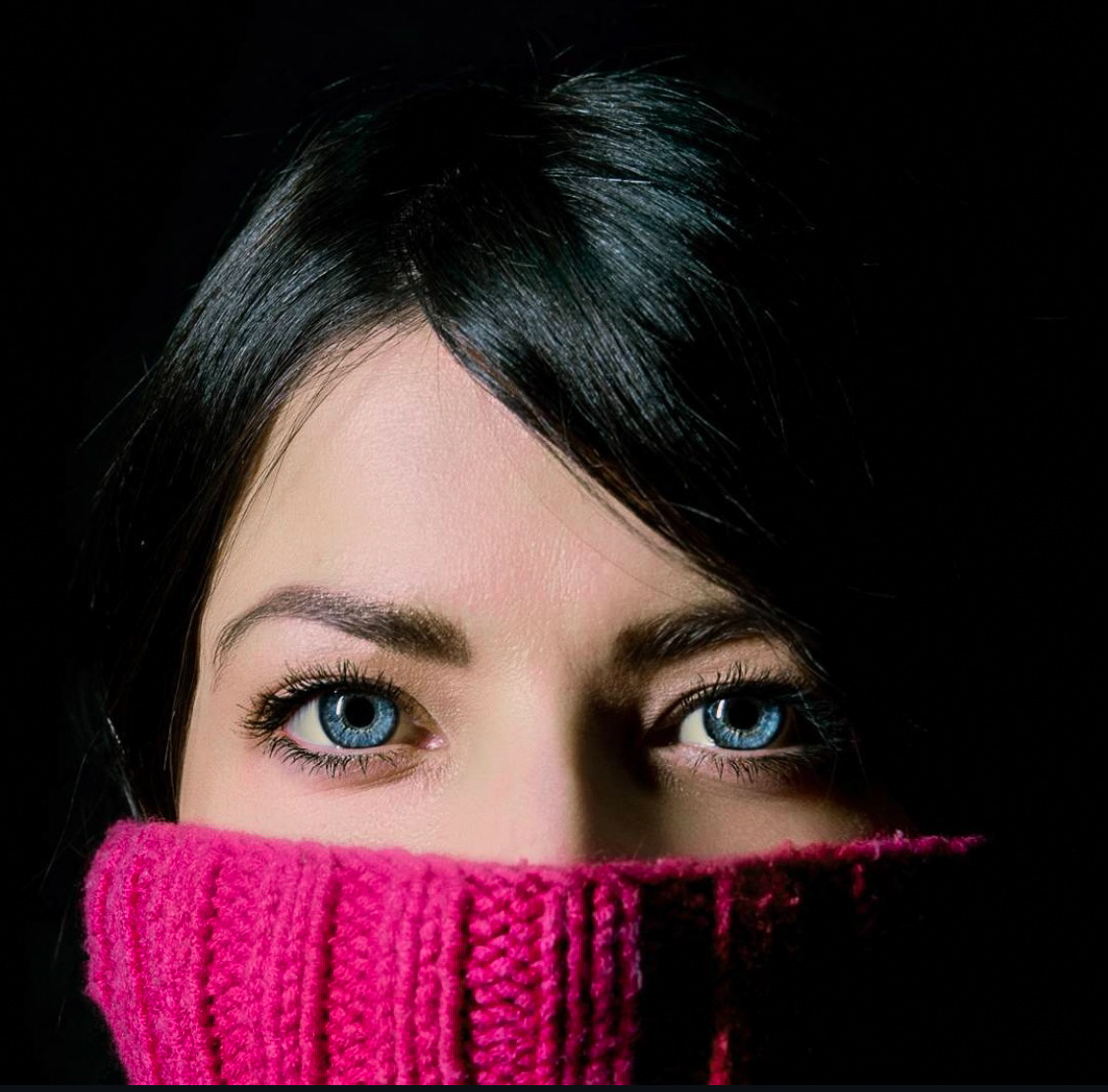 Close-up of a woman's eyes, framed by a pink sweater pulled up to her nose, with dramatic lighting enhancing her piercing gaze