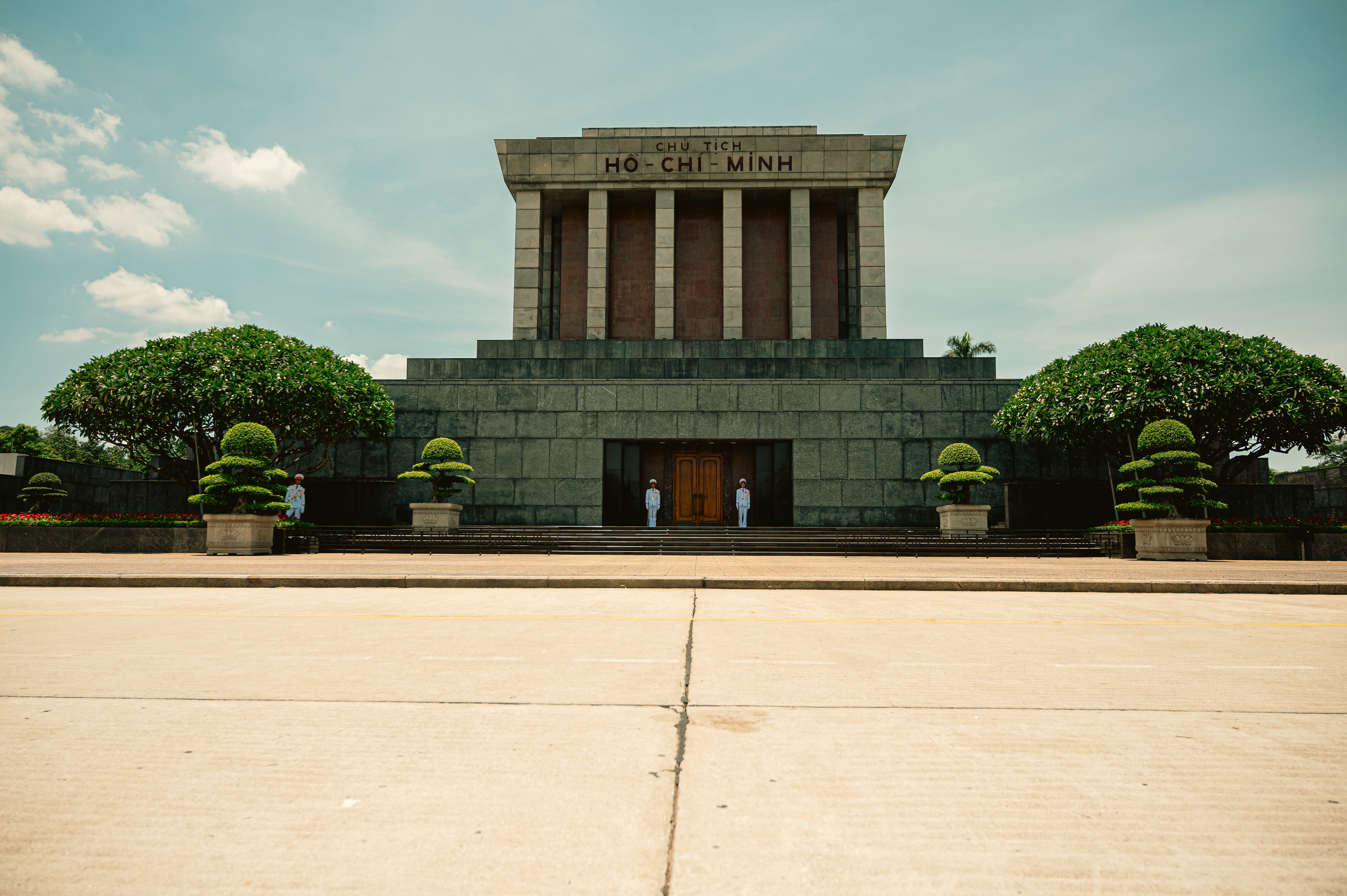 The grand facade of the Ho Chi Minh Mausoleum in Hanoi, Vietnam, framed by manicured gardens and blue skies, with ceremonial guards at attention.