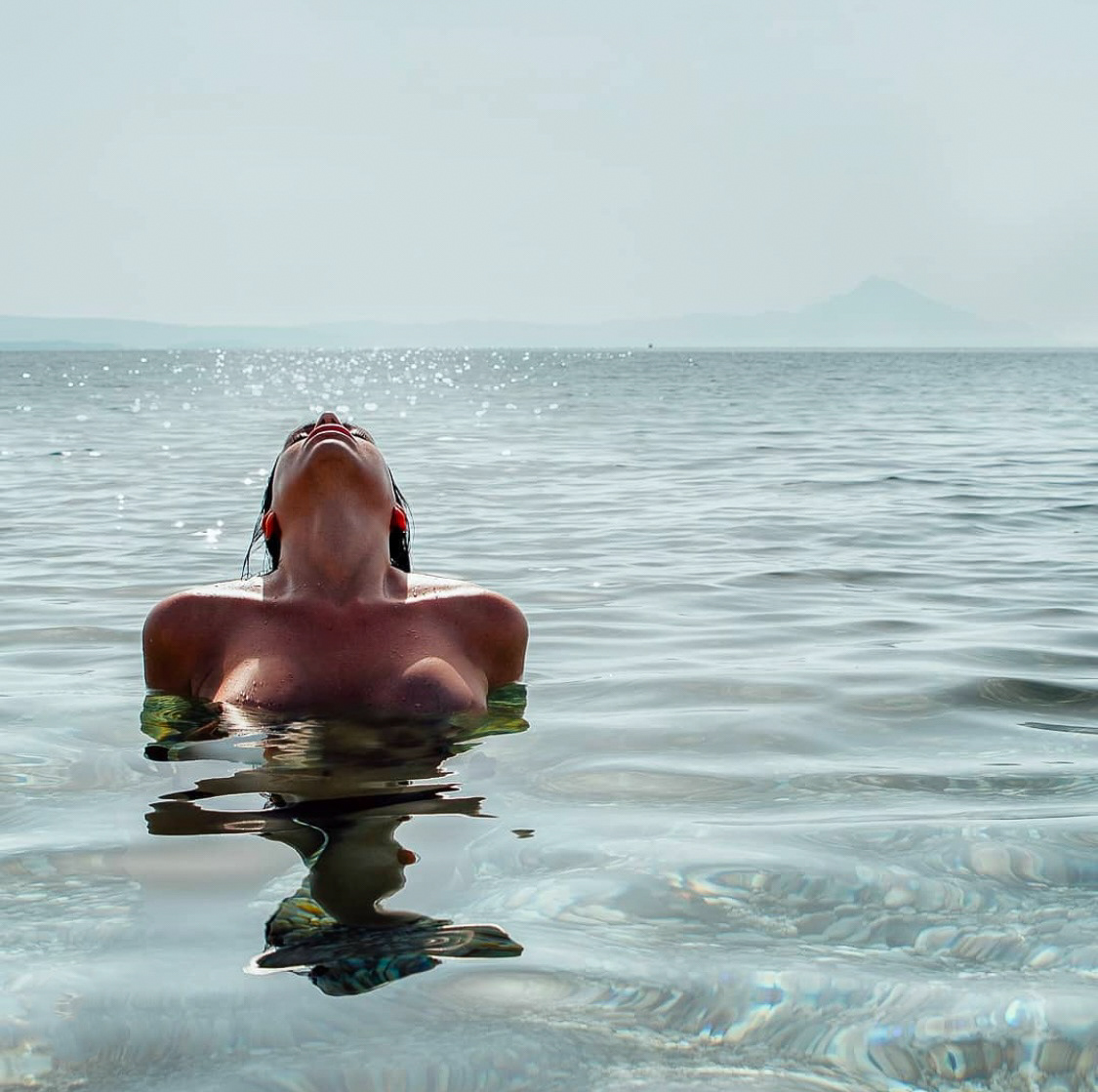 A woman emerging from calm, clear water, tilting her head back as sunlight sparkles on the water's surface.