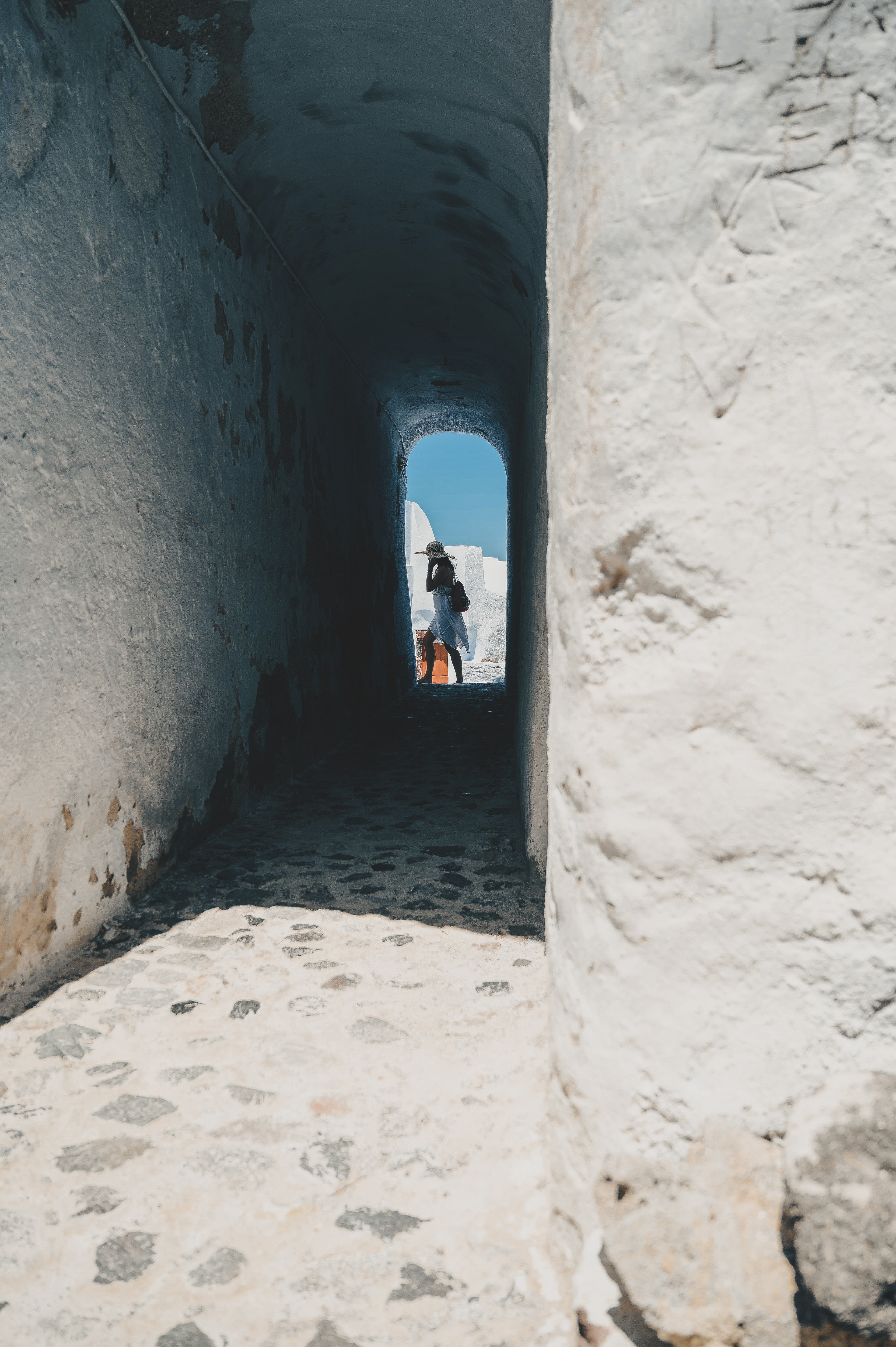 A bright outdoor scene showing a narrow passage with white-washed walls and cobblestone flooring leading to a sunny opening with a woman wearing a wide-brimmed hat in the distance.
