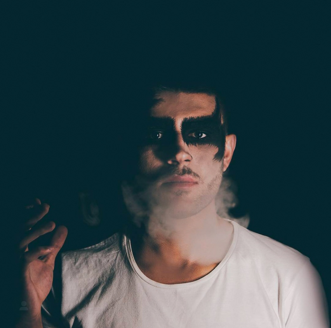 A man with black face paint resembling a mask, exhaling smoke in an enigmatic and theatrical setting.