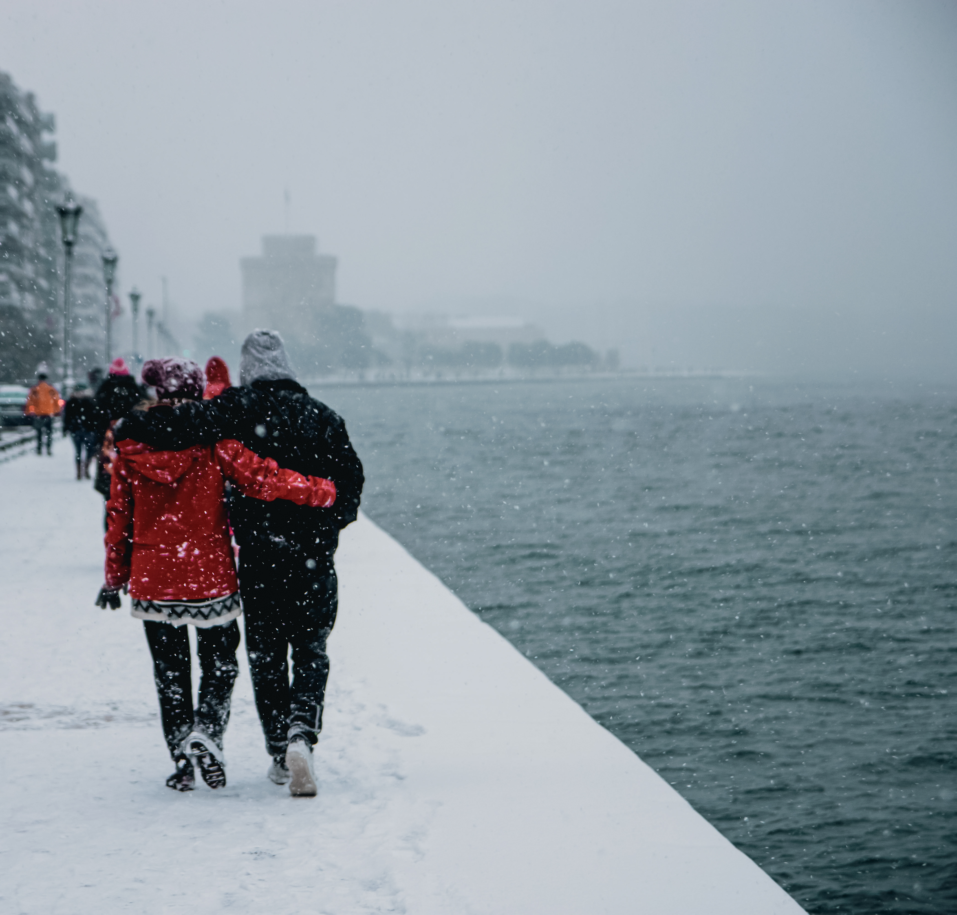 A couple walking arm-in-arm on a snow-covered pathway along a waterfront during snowfall.