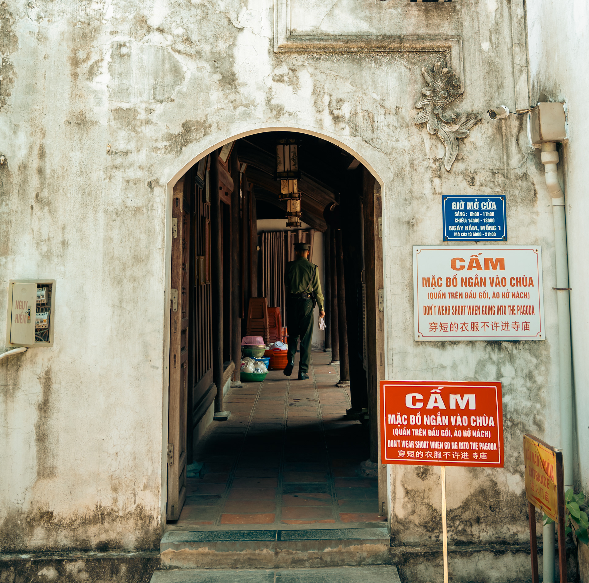A worn temple entrance in Vietnam with signs in multiple languages instructing visitors to dress modestly, featuring a lone guard walking through the corridor.