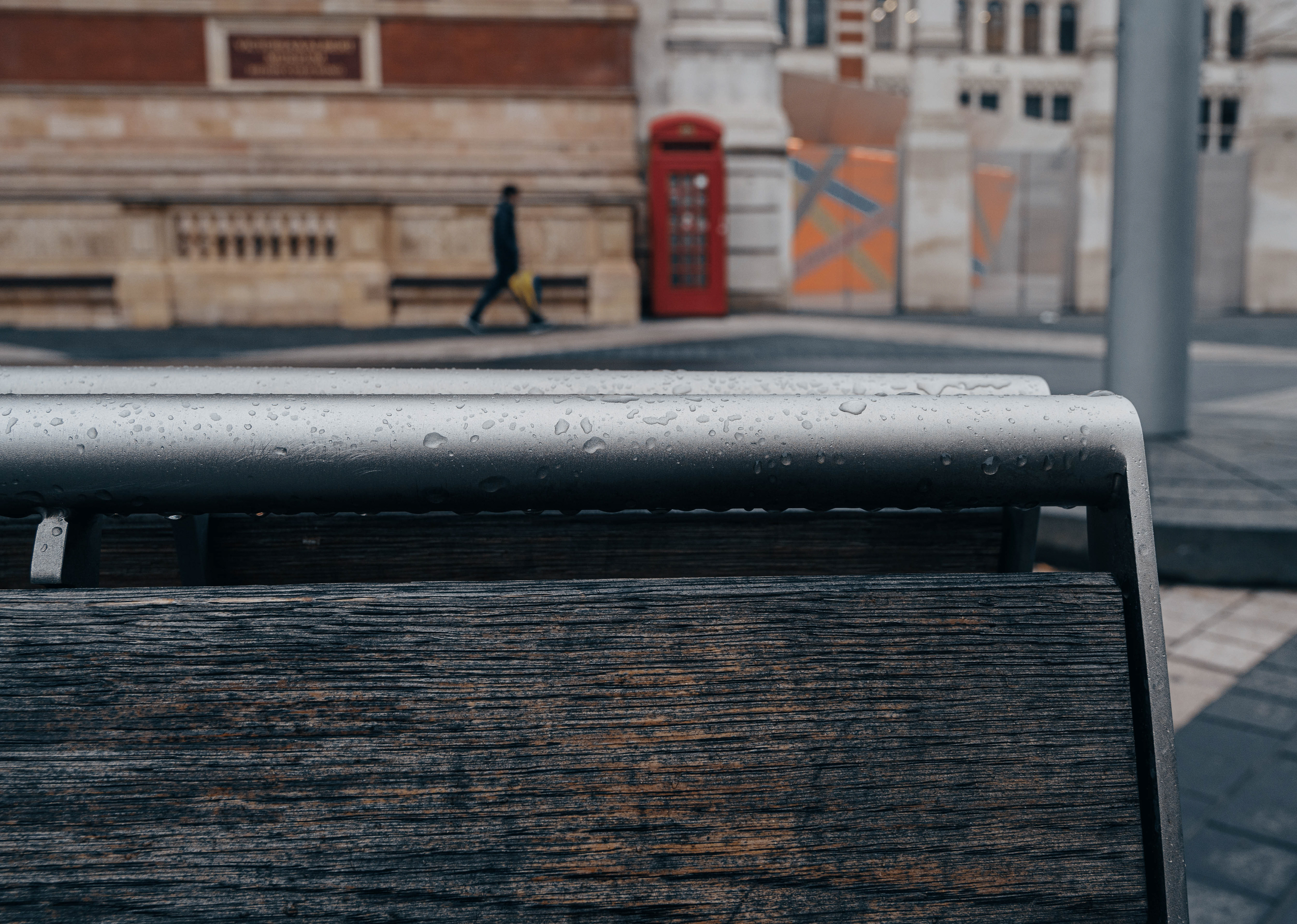 A close-up view of a wooden and metal bench with raindrops, with a blurred background of a red telephone booth and a person walking.