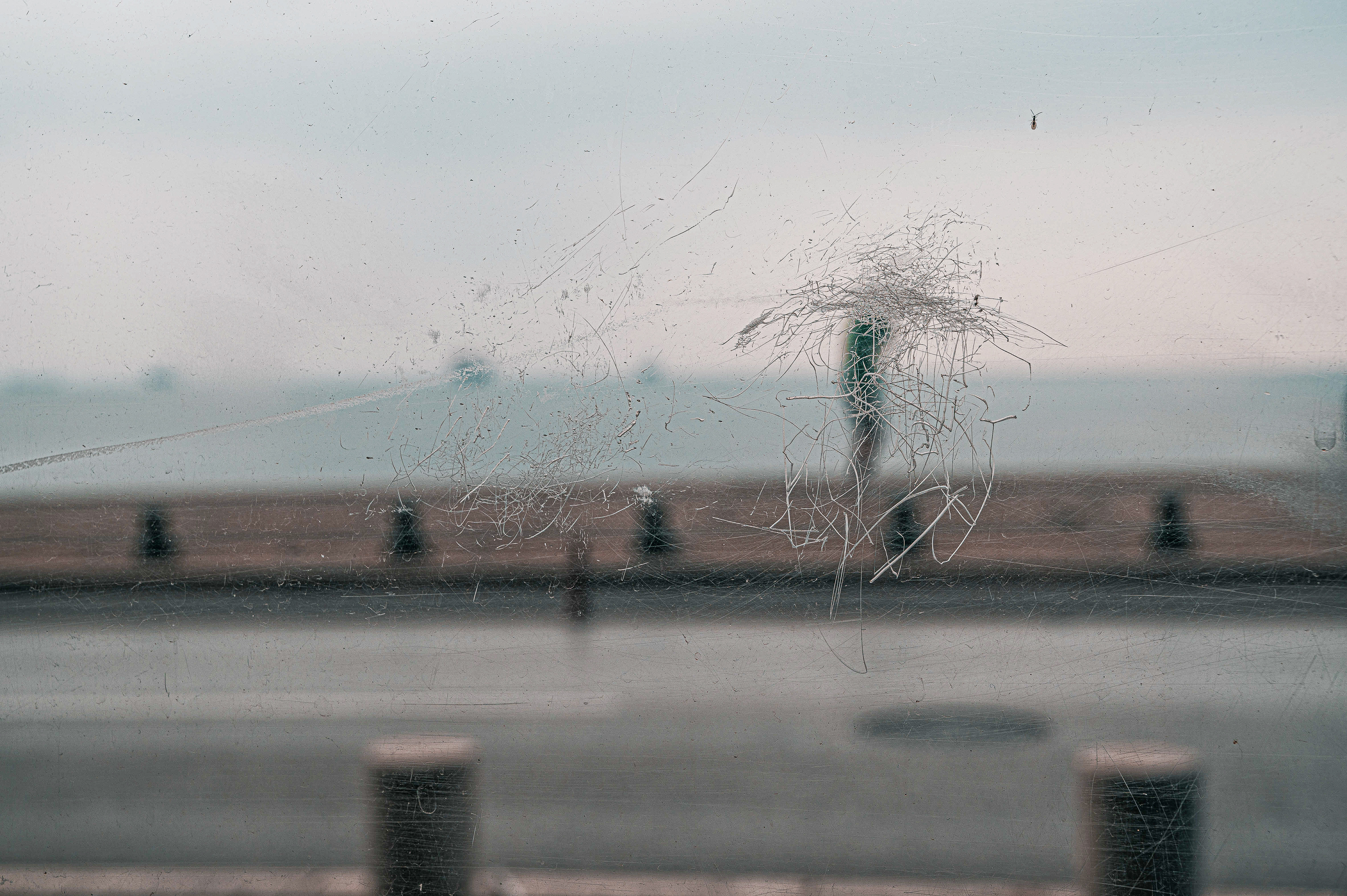 A scratched and slightly opaque window revealing a blurred view of a boardwalk and ocean in the background on a cloudy day.