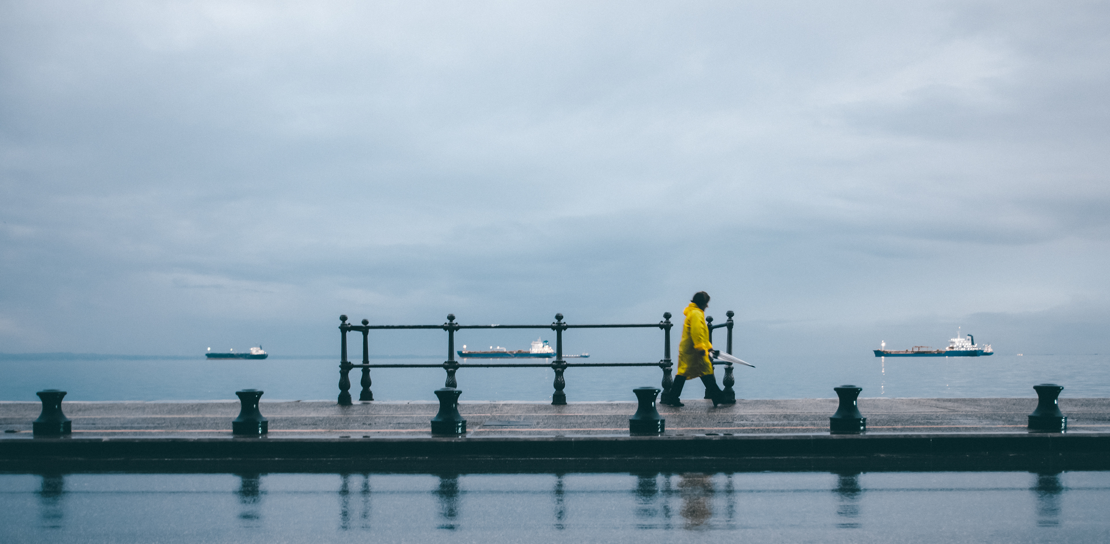 A serene waterfront scene under an overcast sky, featuring a person in a bright yellow raincoat walking along a pier with an umbrella in hand. In the distance, several ships are anchored on the calm sea, framed by the simple black railings of the pier.