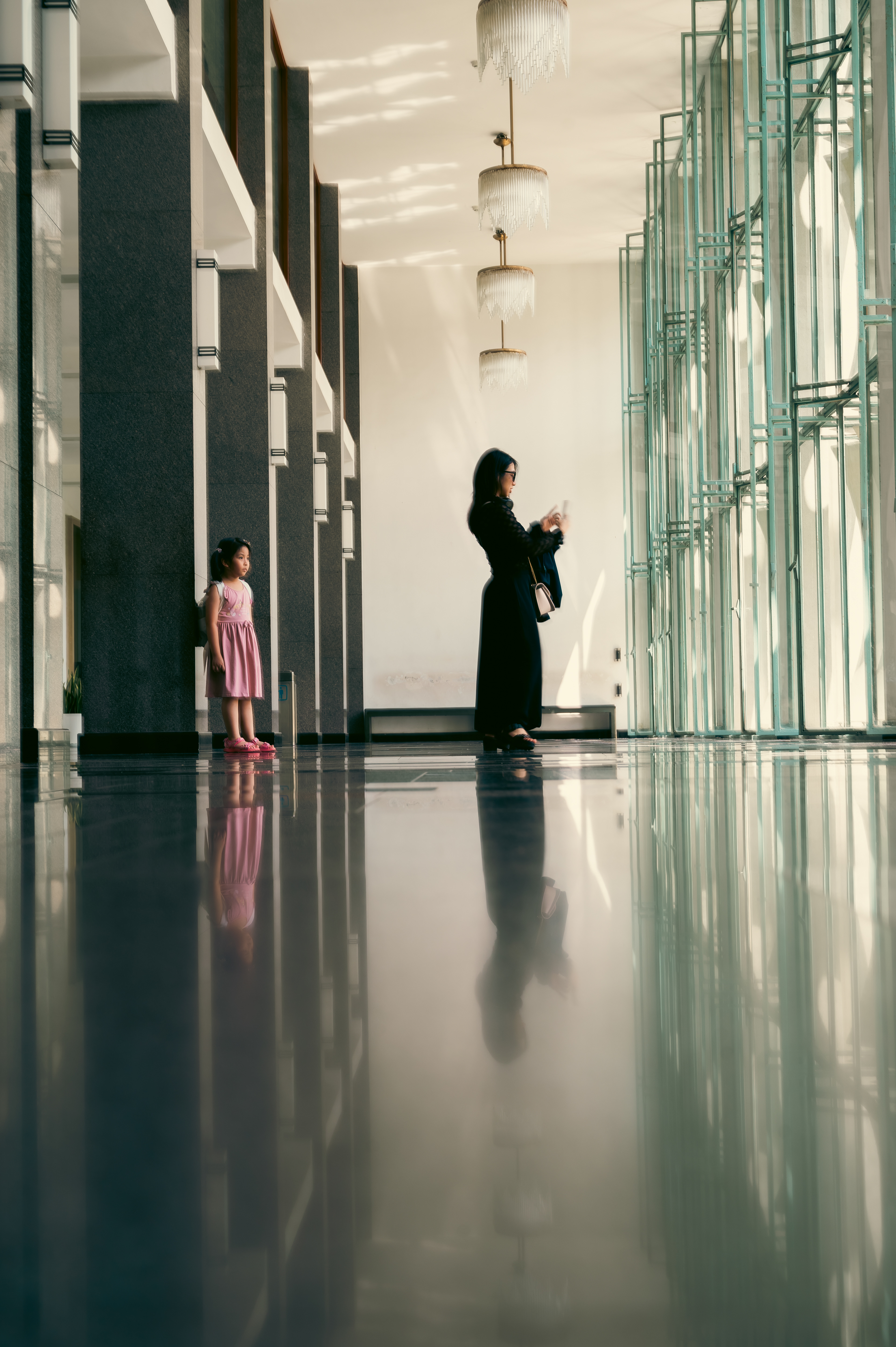 A woman and a young girl stand in a sleek, sunlit hall with reflective floors, framed by geometric glass panels and elegant chandeliers.\