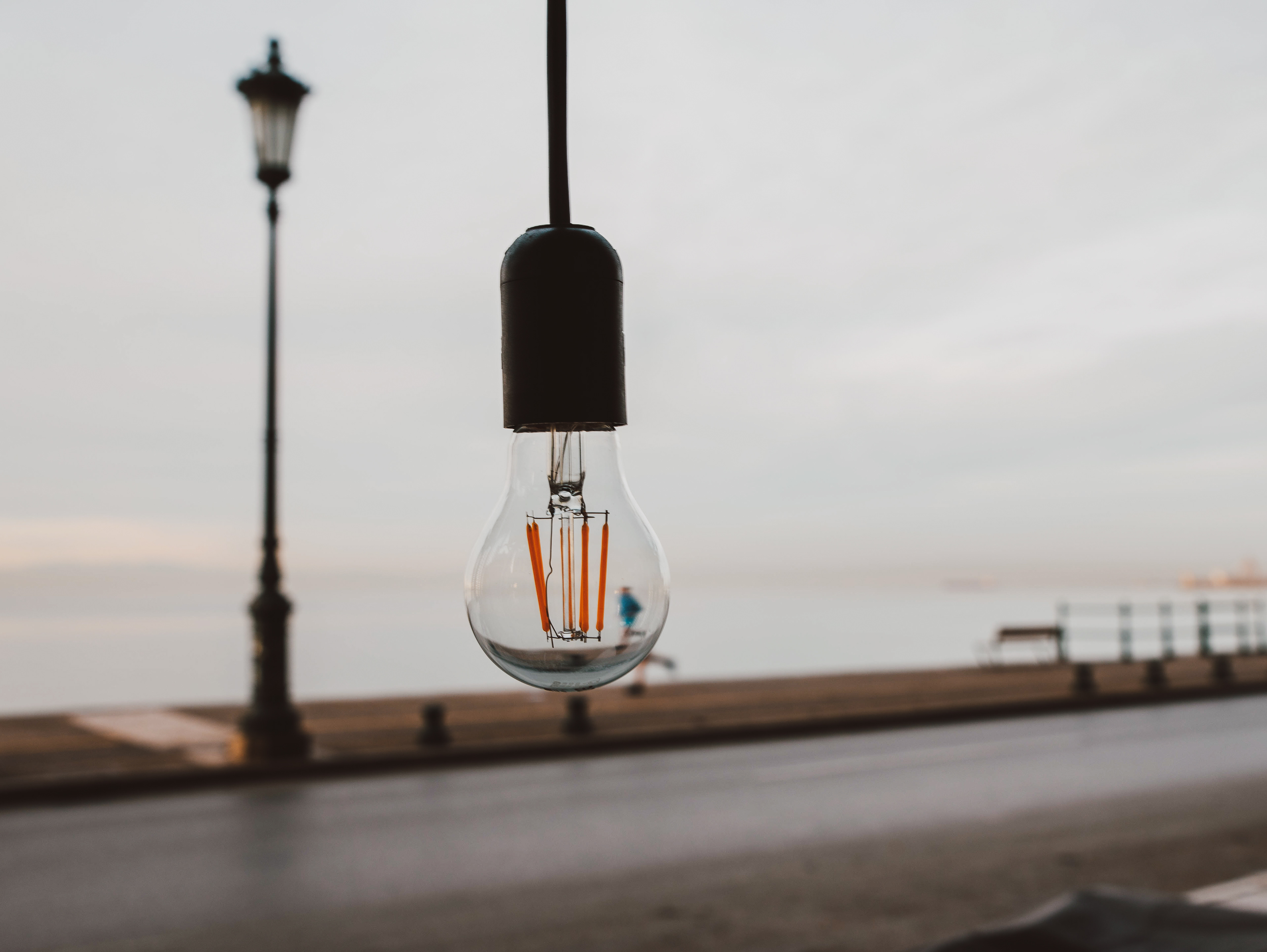 A lightbulb hanging in focus, capturing a softly blurred seaside promenade with streetlamps in the background.