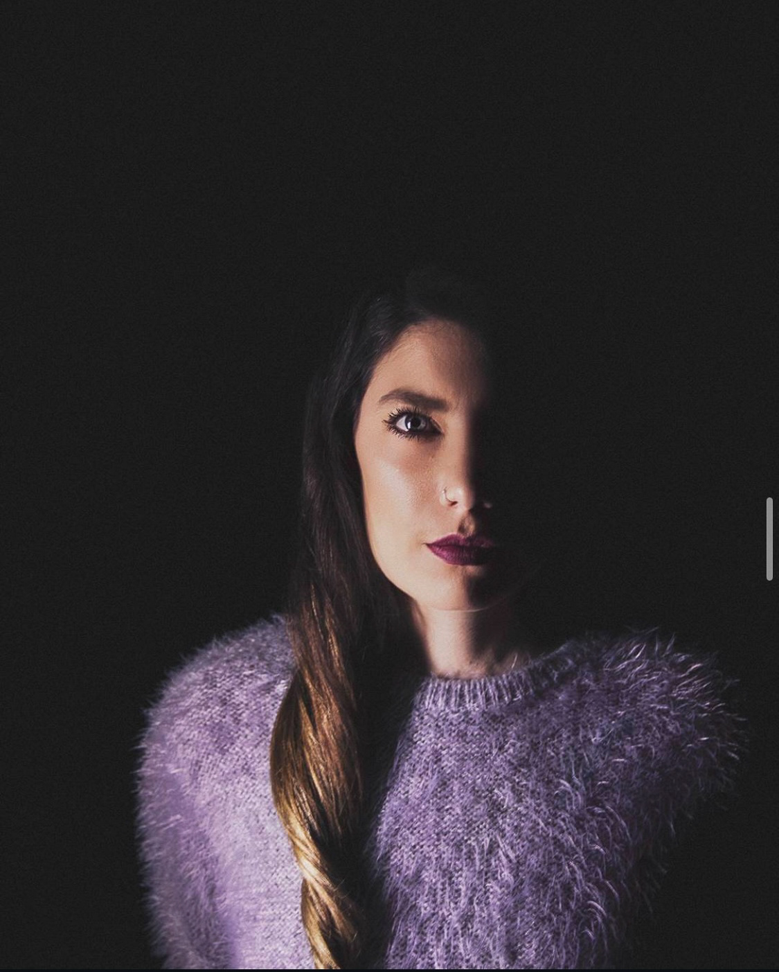 "A dramatic portrait of a woman with long hair wearing a soft purple sweater, half her face illuminated in low-key lighting.