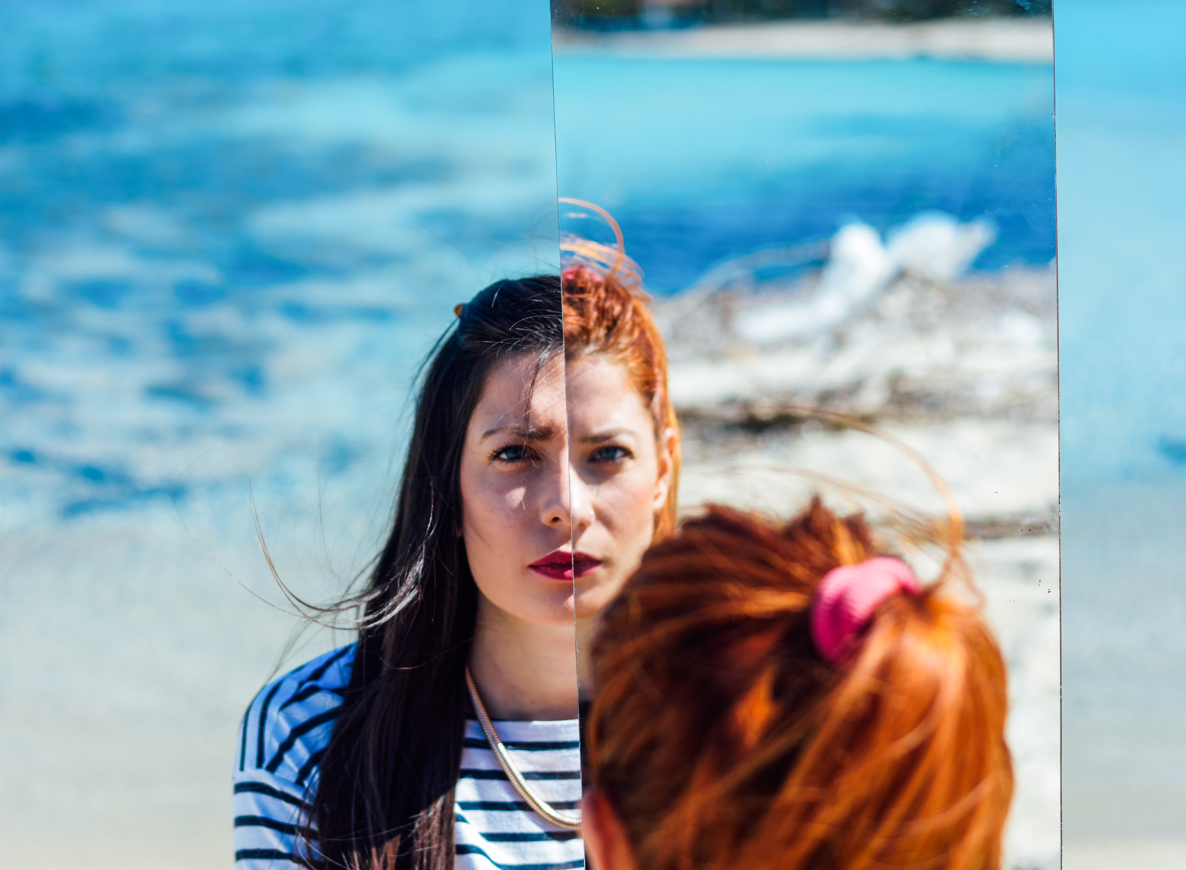 Portrait of twin sisters at the beach, captured through a mirror that splits their reflections, creating a striking visual effect. The vibrant blue ocean and sandy shoreline provide a serene backdrop, emphasizing their connection and individuality.