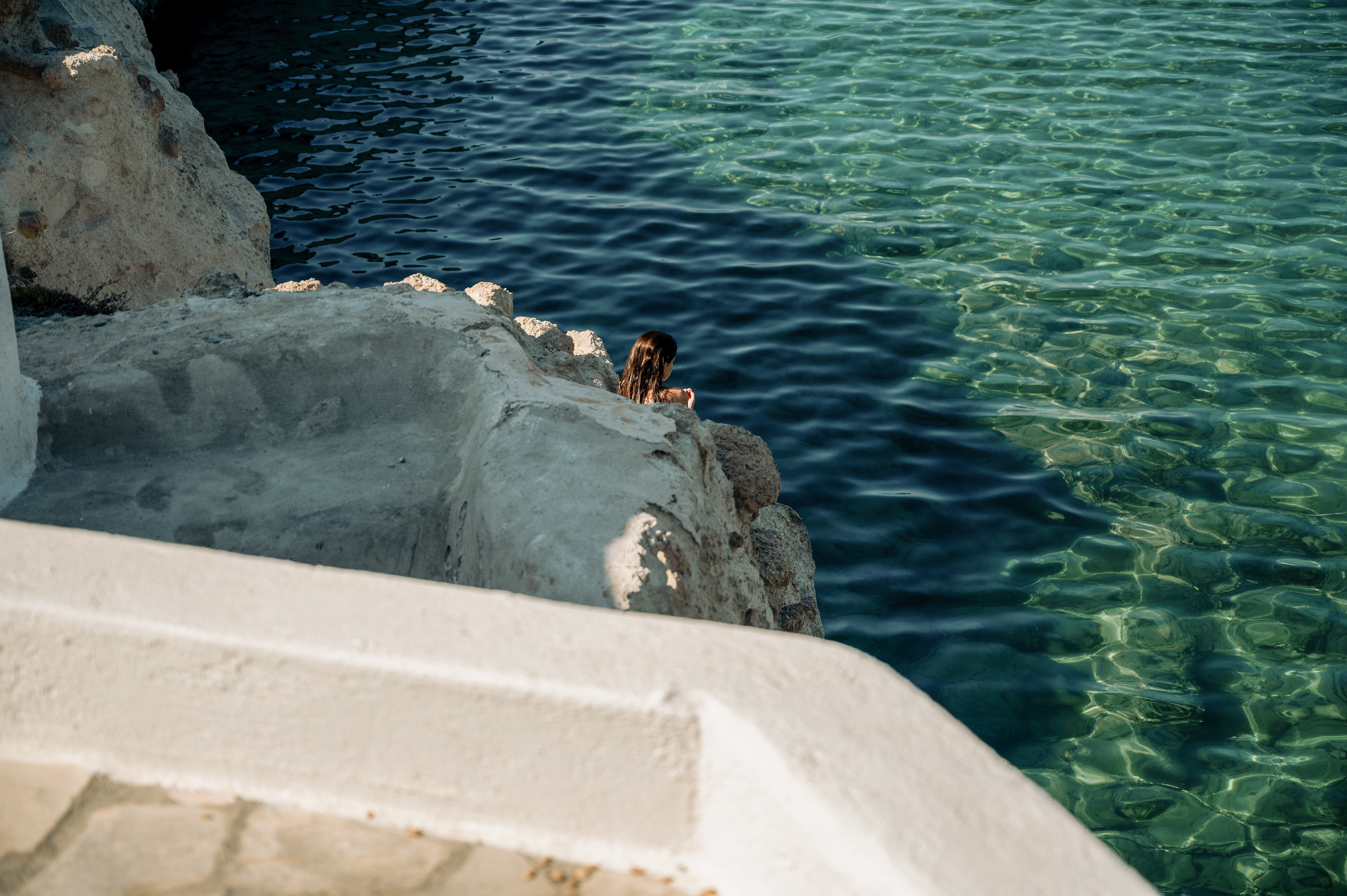 A woman sits near a rugged white structure, gazing at the crystal-clear turquoise water below, creating a tranquil, coastal vibe..