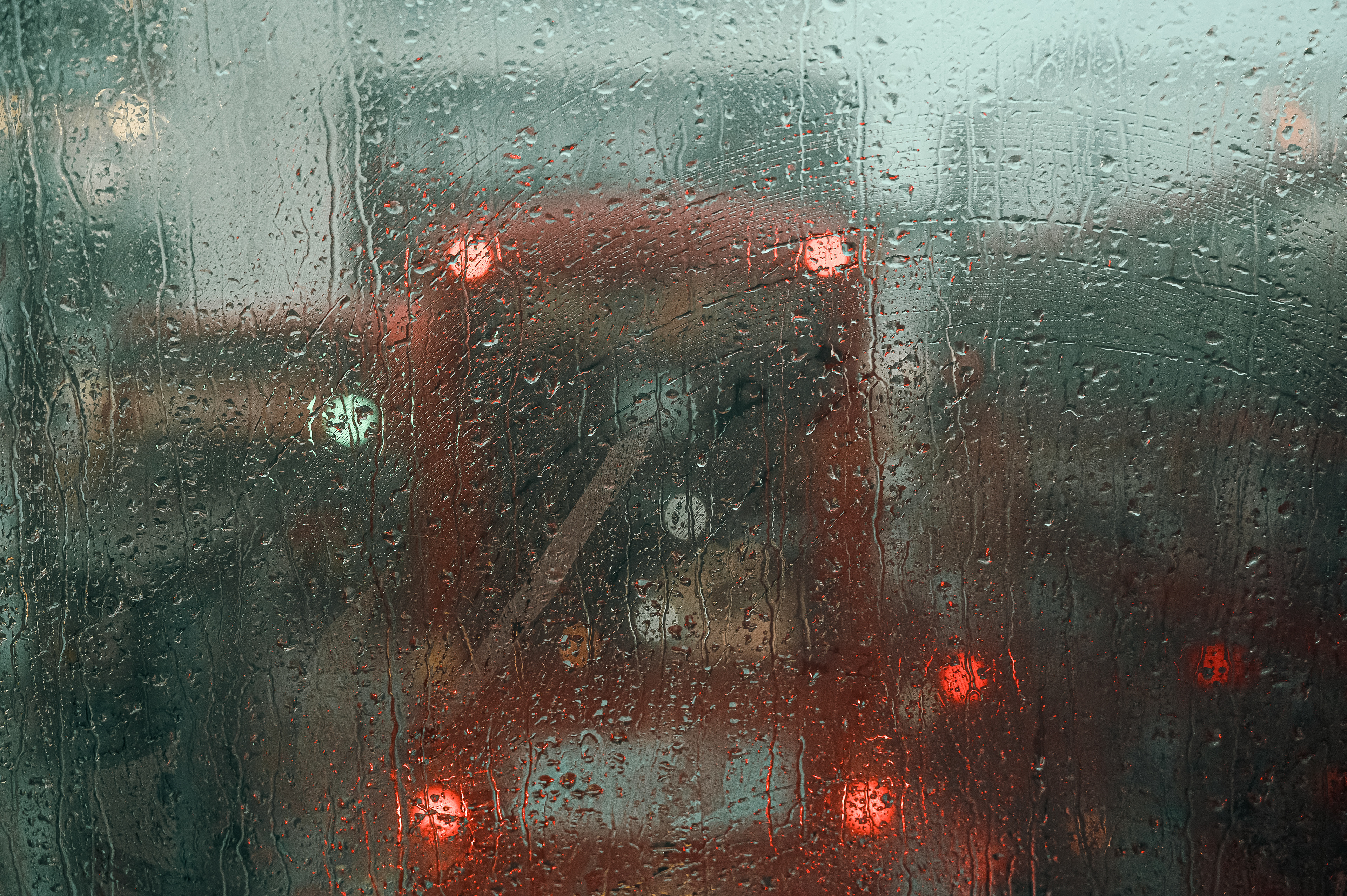 A rainy street scene captured through a heavily water-streaked window, with blurred red buses and brake lights visible in the background.