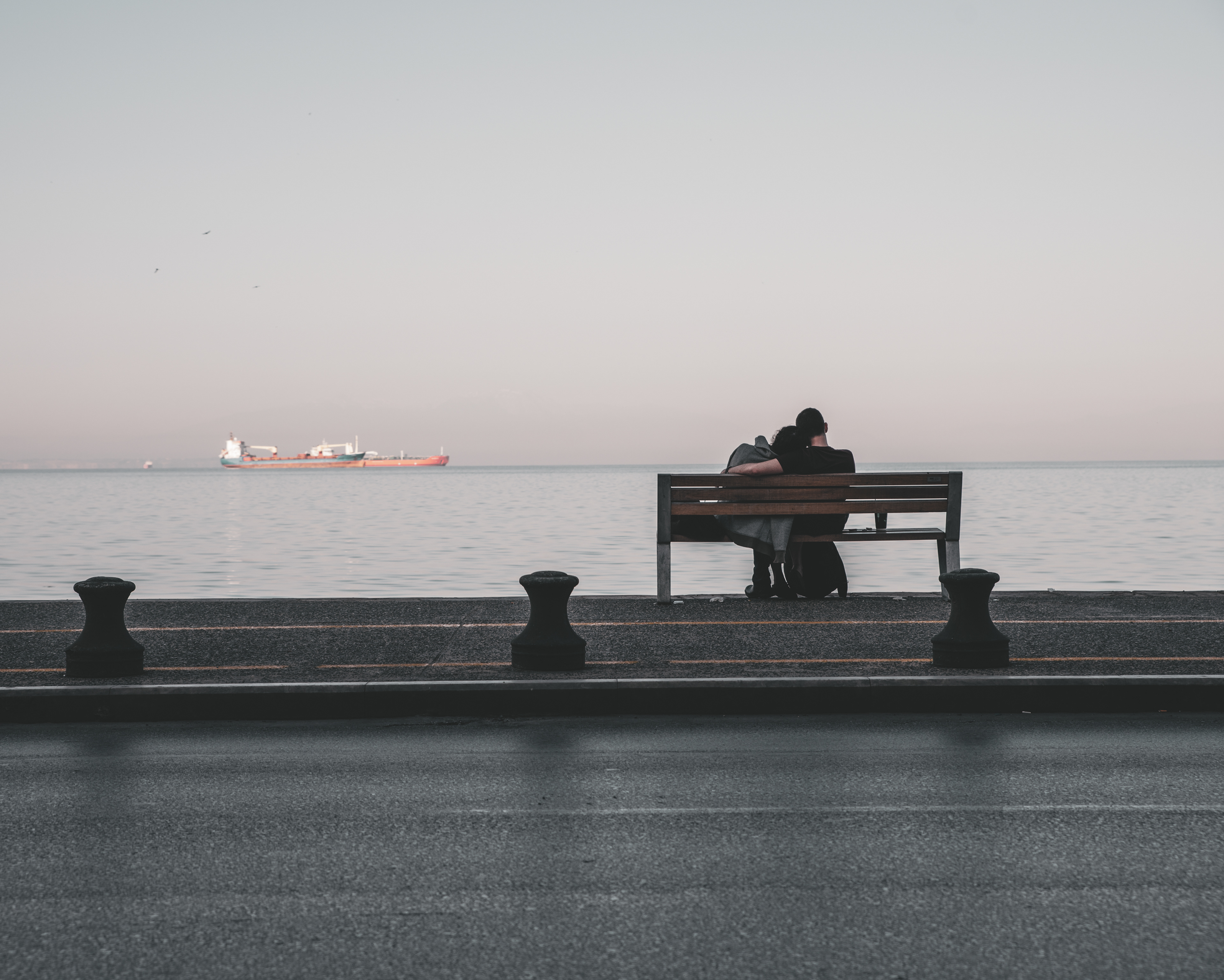 A couple sitting on a bench facing the sea, with a large ship in the distance, under a serene evening sky