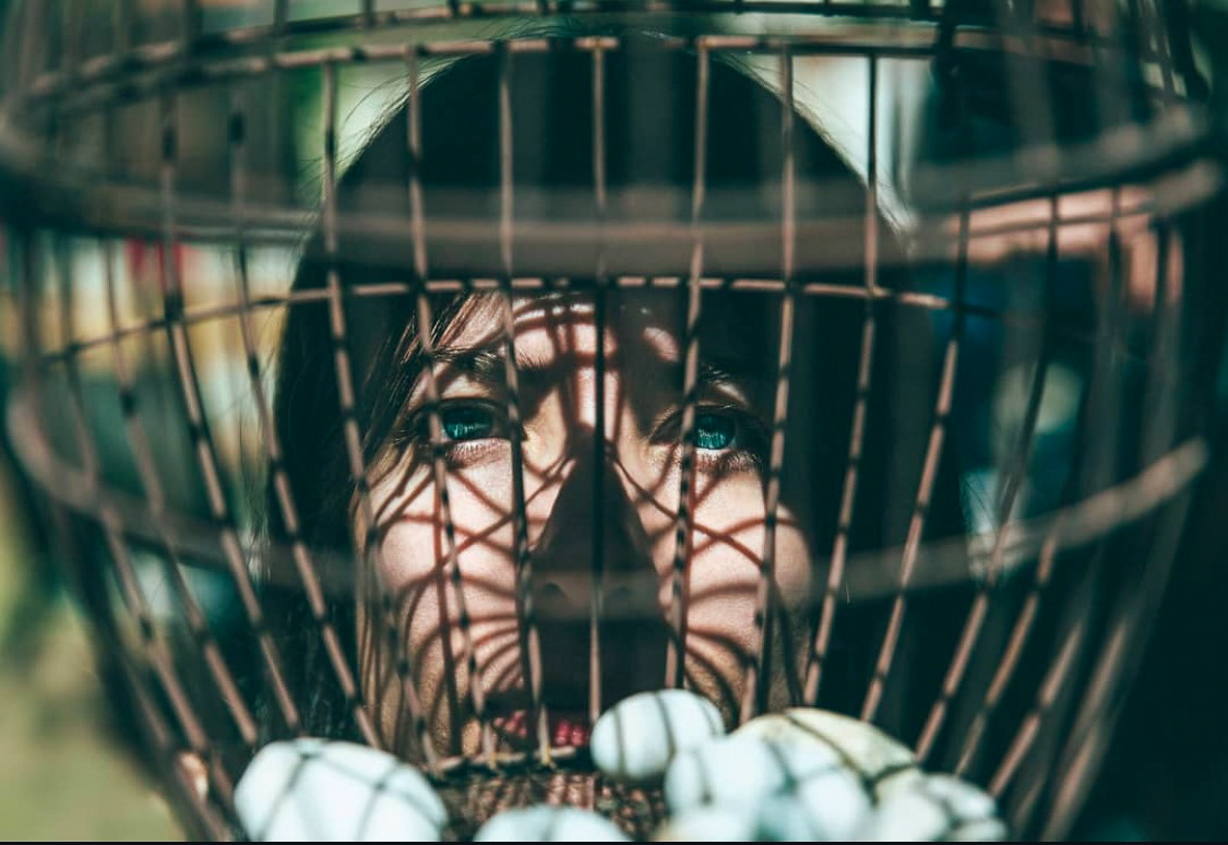 A woman framed behind the shadowy bars of a circular cage-like structure, her intense blue eyes piercing through the shadows.