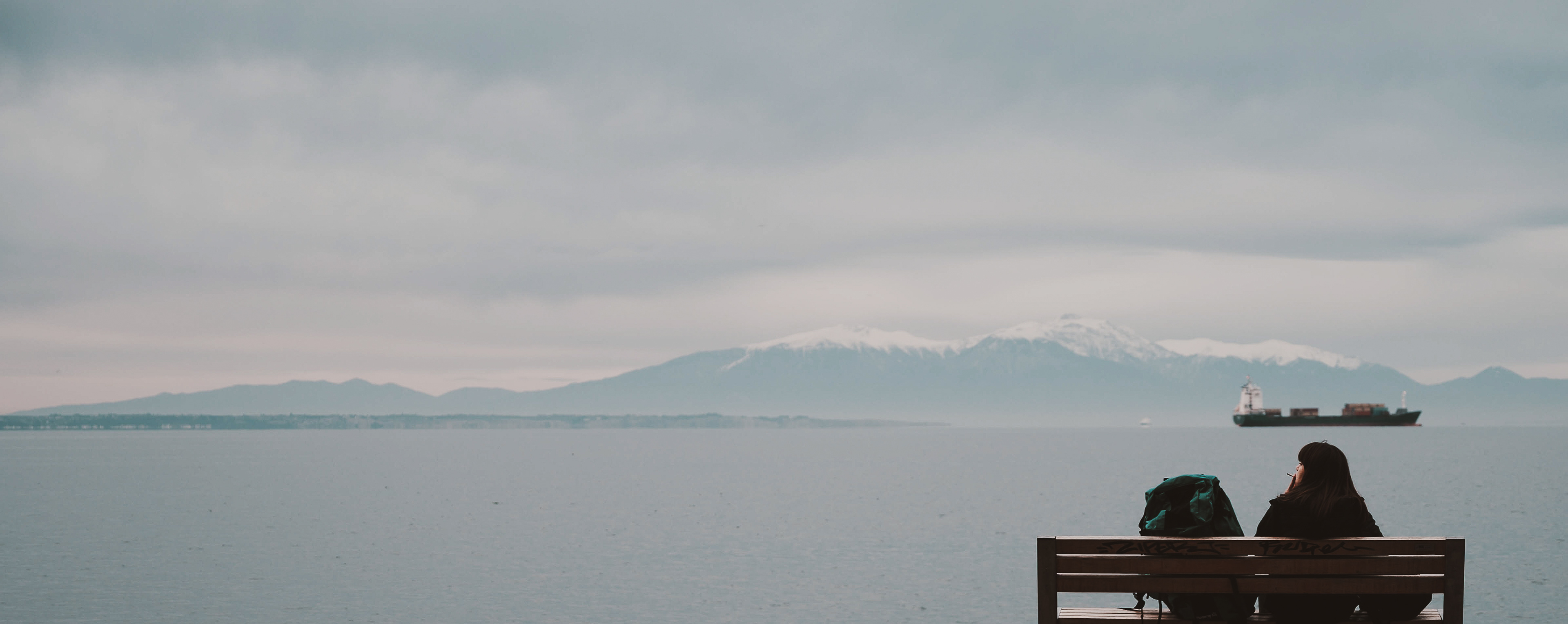 A panoramic view of a bench by a calm sea, with two people sitting and a large mountain range in the distant background under a cloudy sky.