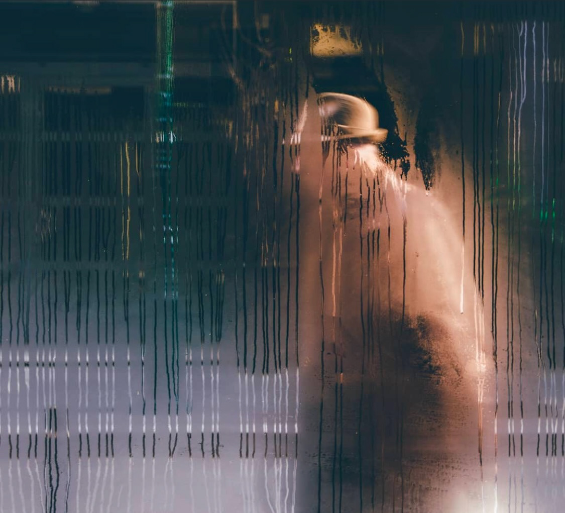 A hazy silhouette of a person wearing a hat, captured through a fogged-up window, creating an atmospheric scene.