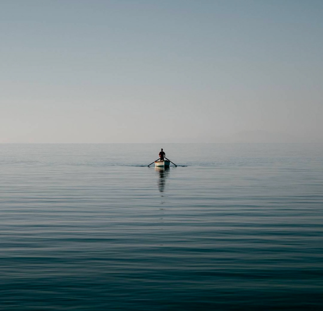 A lone person rows a small boat on a still, expansive sea under a vast, pale sky.