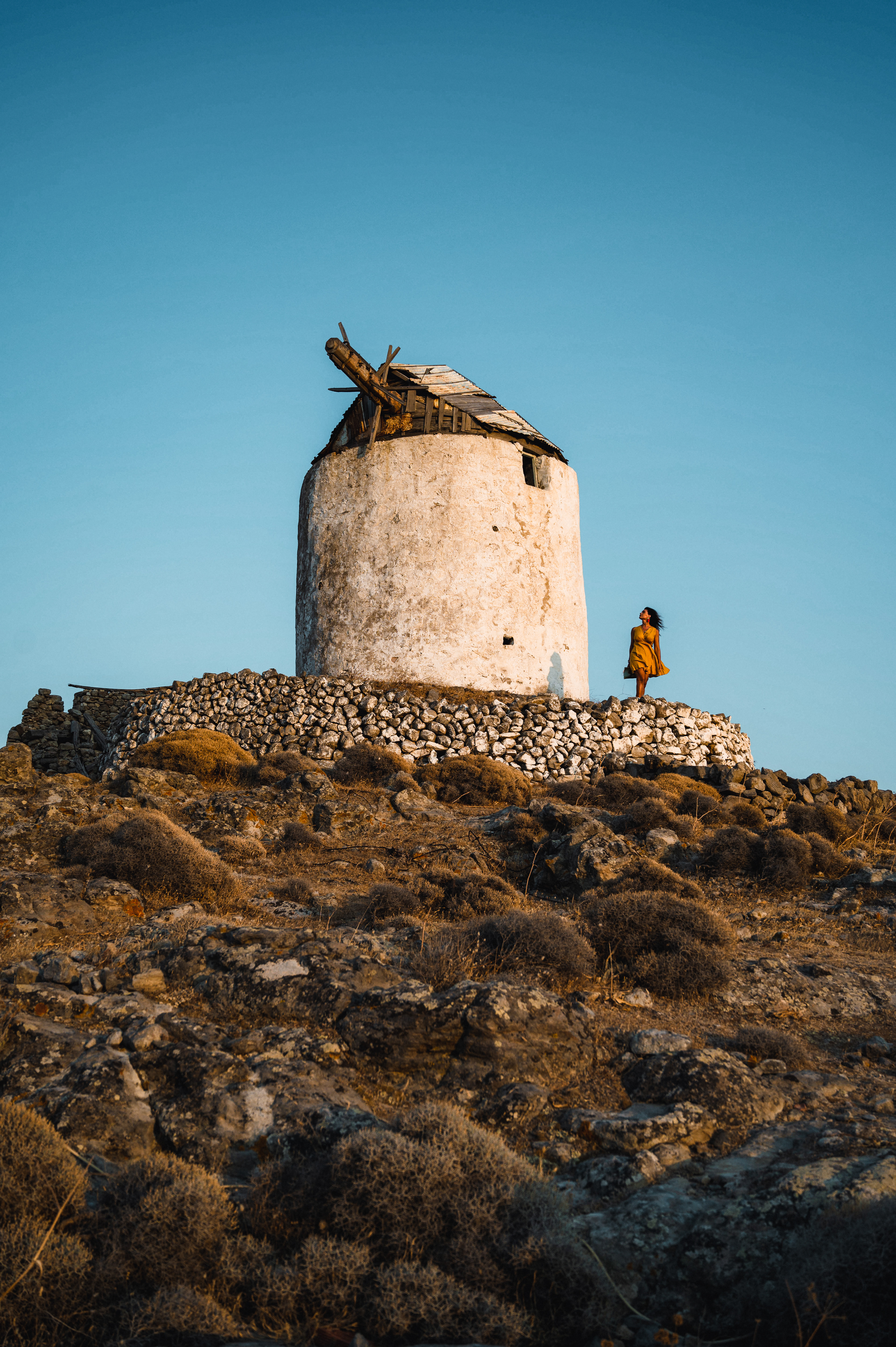 A solitary, rustic windmill atop a rocky hill under a clear blue sky, with a woman in a flowing yellow dress standing nearby, evoking a sense of timelessness.