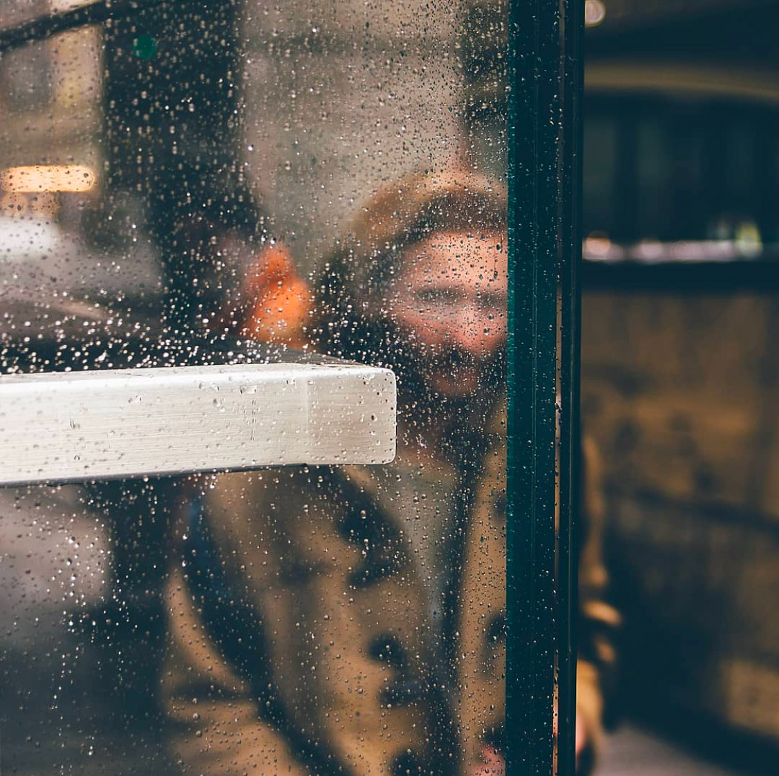 A blurred figure visible through a glass pane covered in raindrops, evoking a contemplative, moody ambiance.