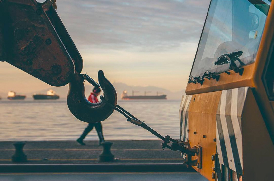 A close-up view of a heavy-duty crane hook against a serene seaside background with ships docked under the golden hour light.