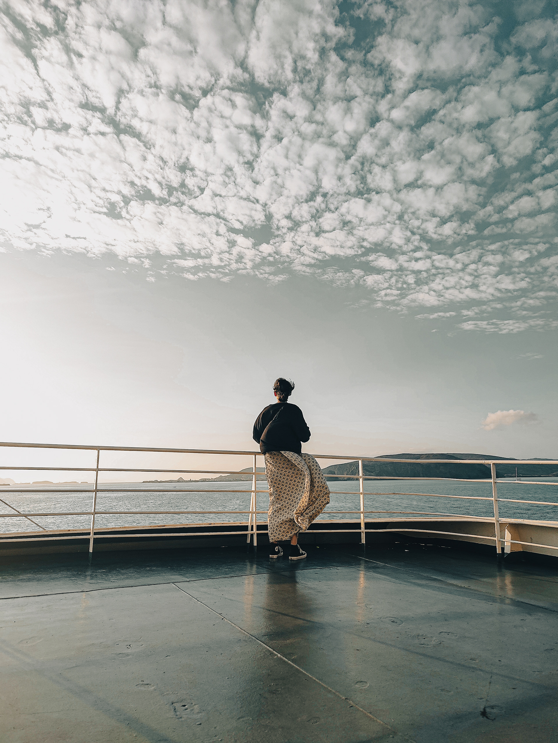 A serene photo of a woman standing on a ferry deck, gazing at the vast ocean and cloudy sky, capturing a peaceful moment of solitude