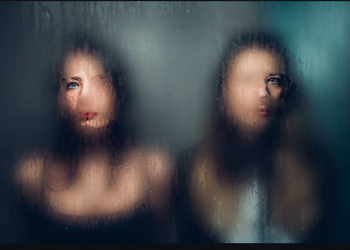 Two women standing behind a foggy, steamy glass, their faces partially blurred and visible through droplets.