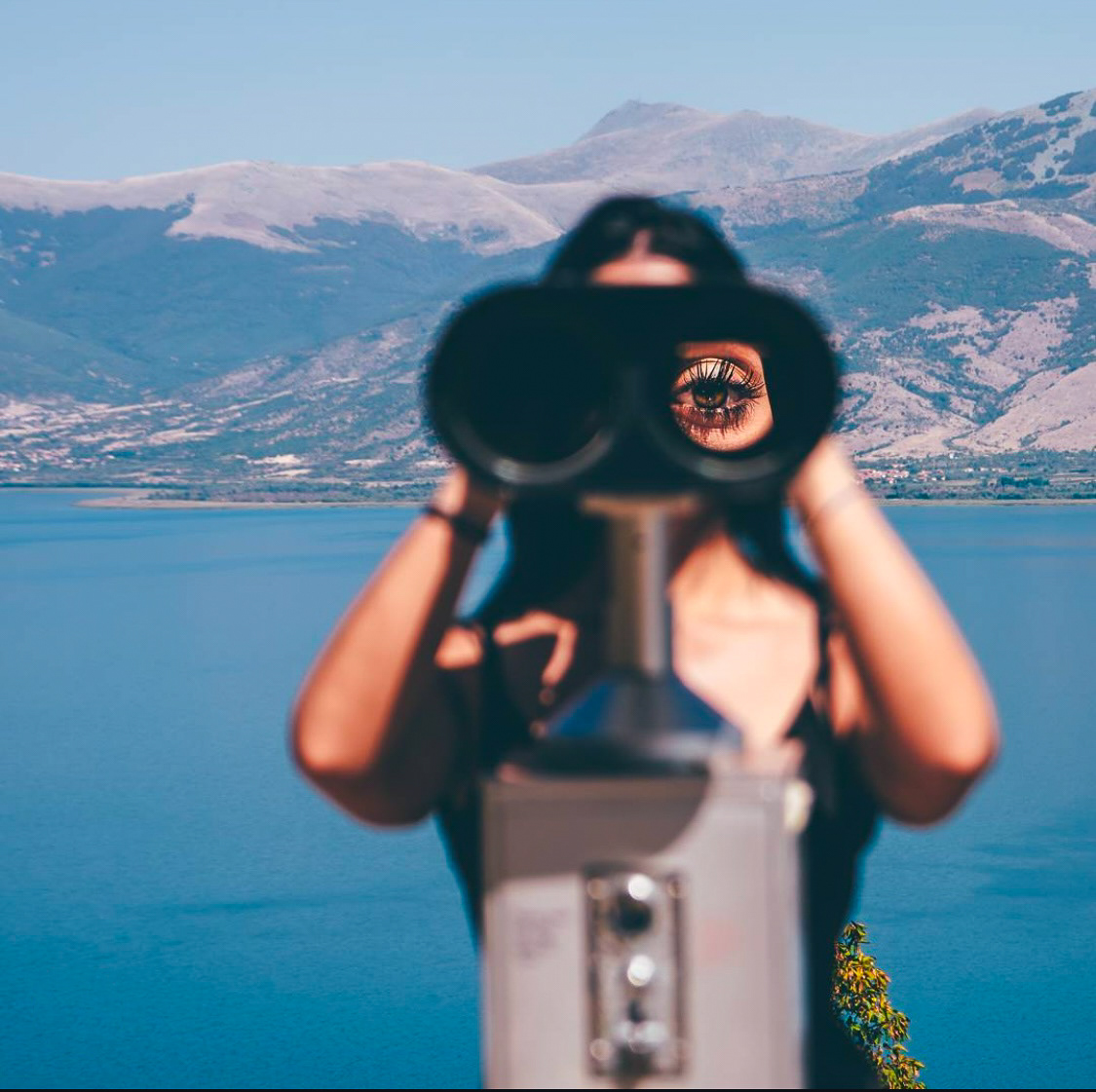 A woman gazing through binoculars with her eye visible in one lens, set against a serene mountain and lake backdrop.