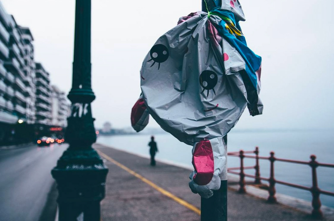 A deflated cartoon-themed balloon tied to a streetlamp post, with a distant figure walking along a coastal promenade in the background.