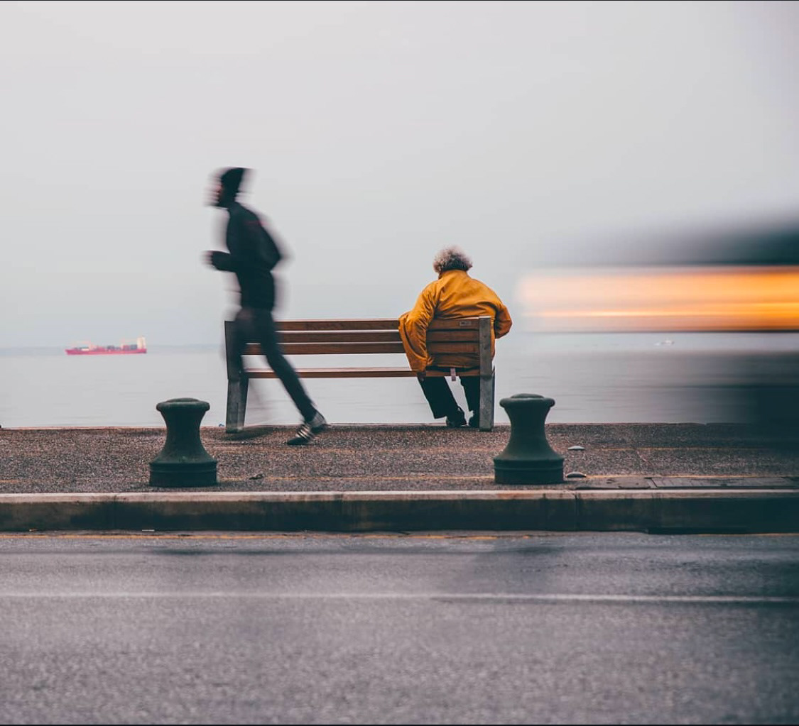 A seated elderly person in a yellow jacket on a bench overlooks a calm sea, while a jogger and a blurred passing object symbolize motion in the foreground.
