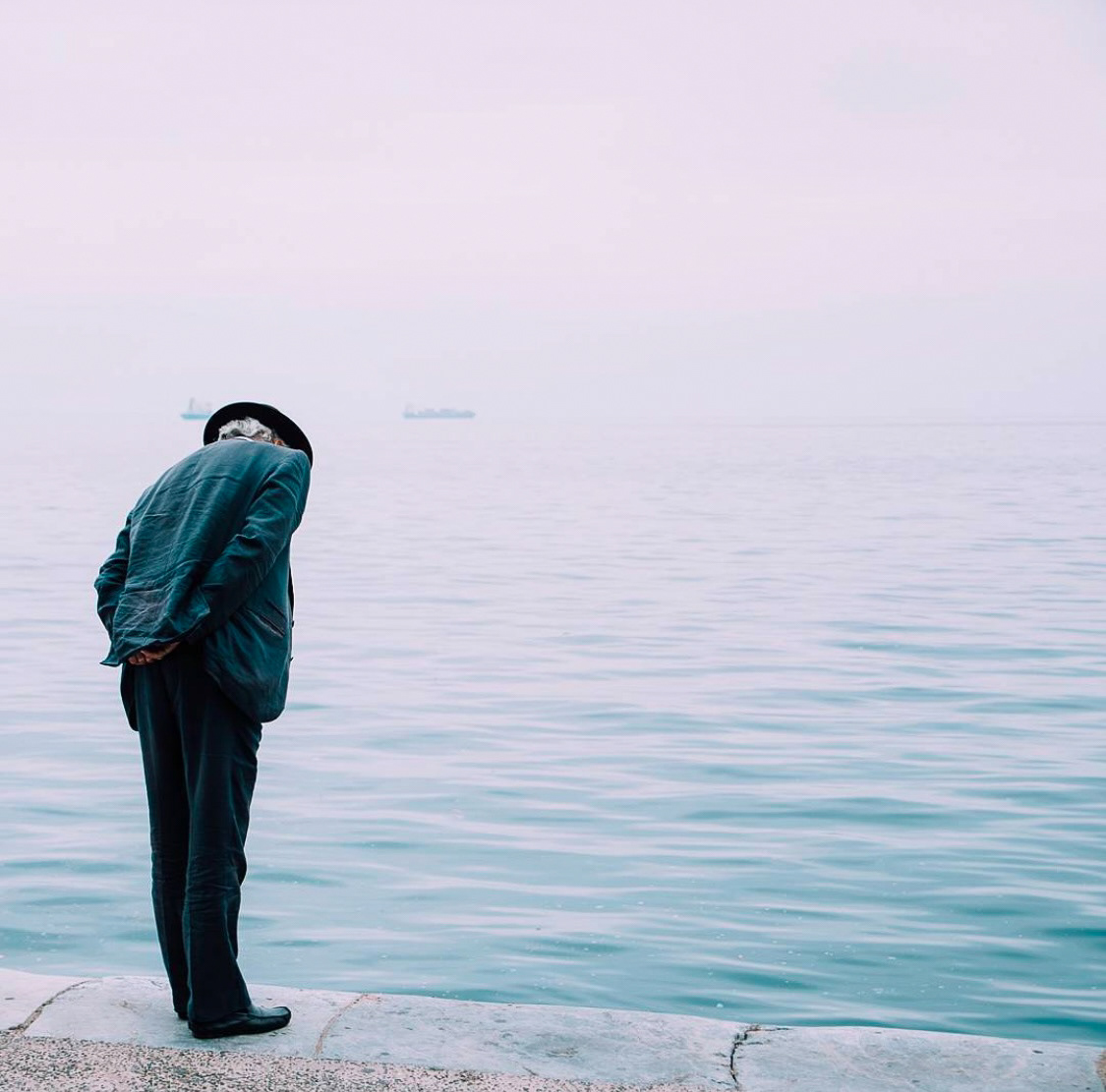 An elderly man in a dark suit and hat gazes contemplatively at the calm waters of a misty seafront.