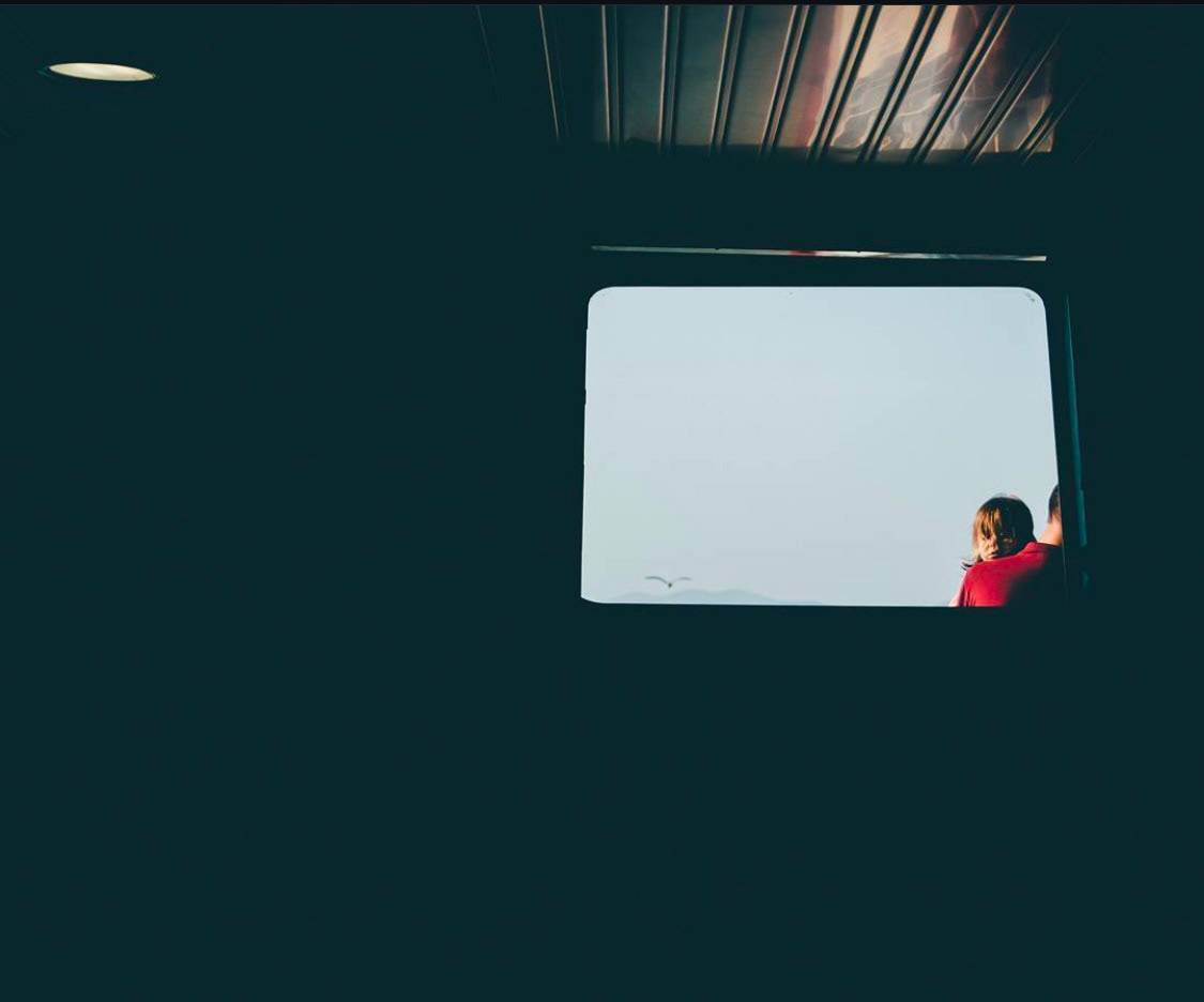 Two figures, possibly a parent and child, seen through a window frame looking out at a serene seascape, suggesting a moment of reflection.