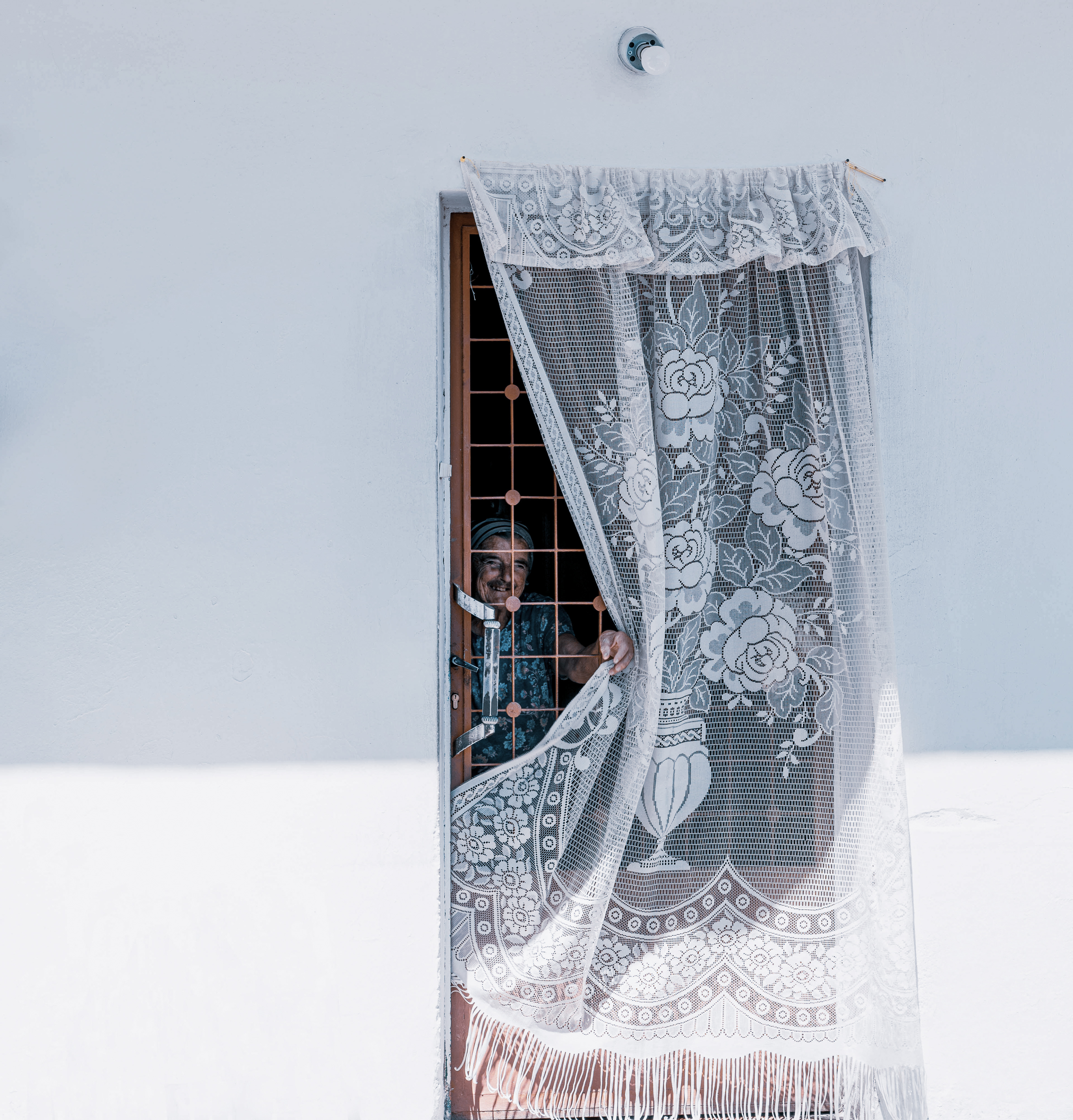 A close-up of a doorway adorned with delicate lace curtains, partially drawn back by an elderly person peeking out, against a white wall background.