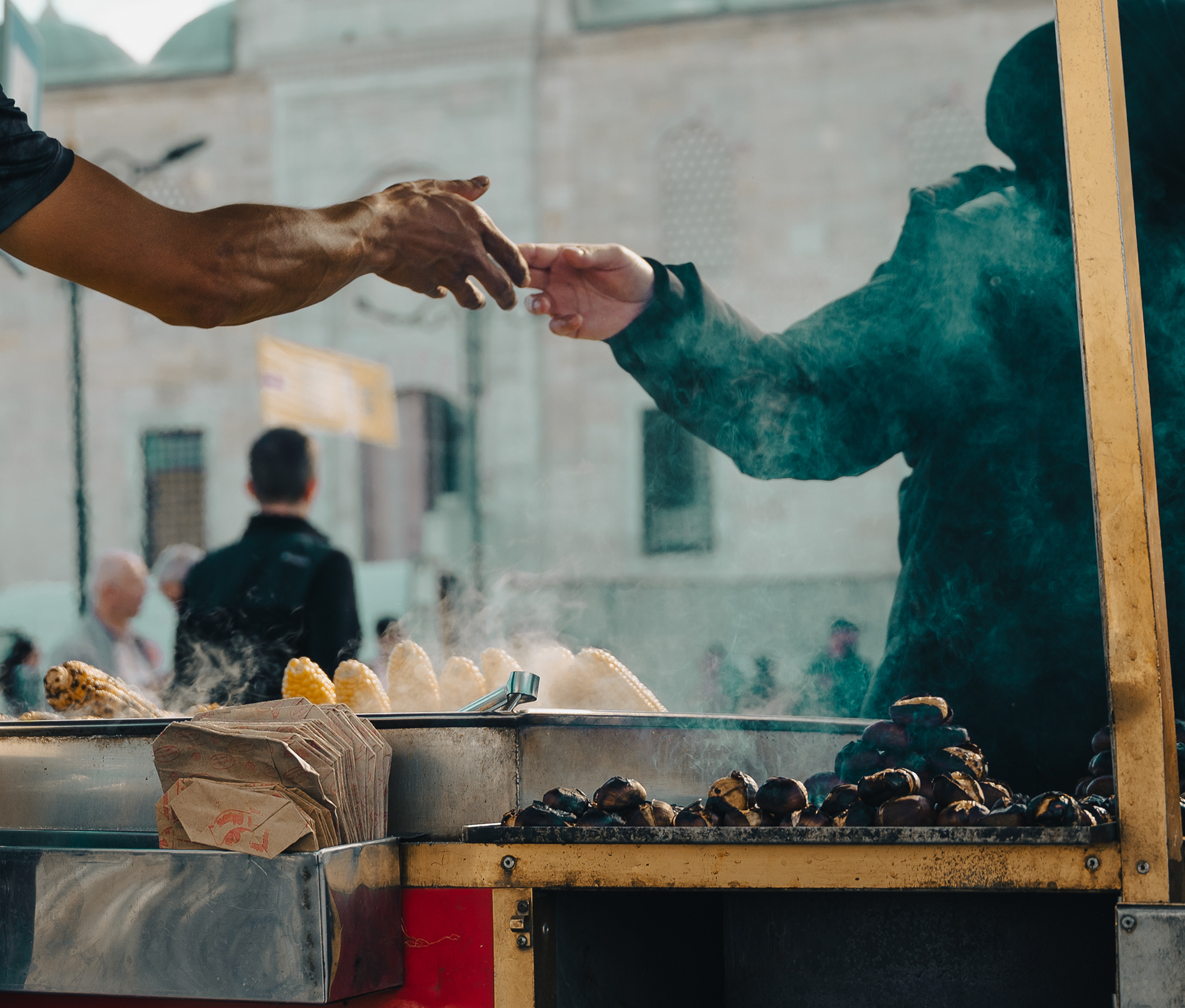 Close-up of hands exchanging money at a street food cart, with steaming roasted chestnuts and corn in the foreground and blurred urban scenery in the background.