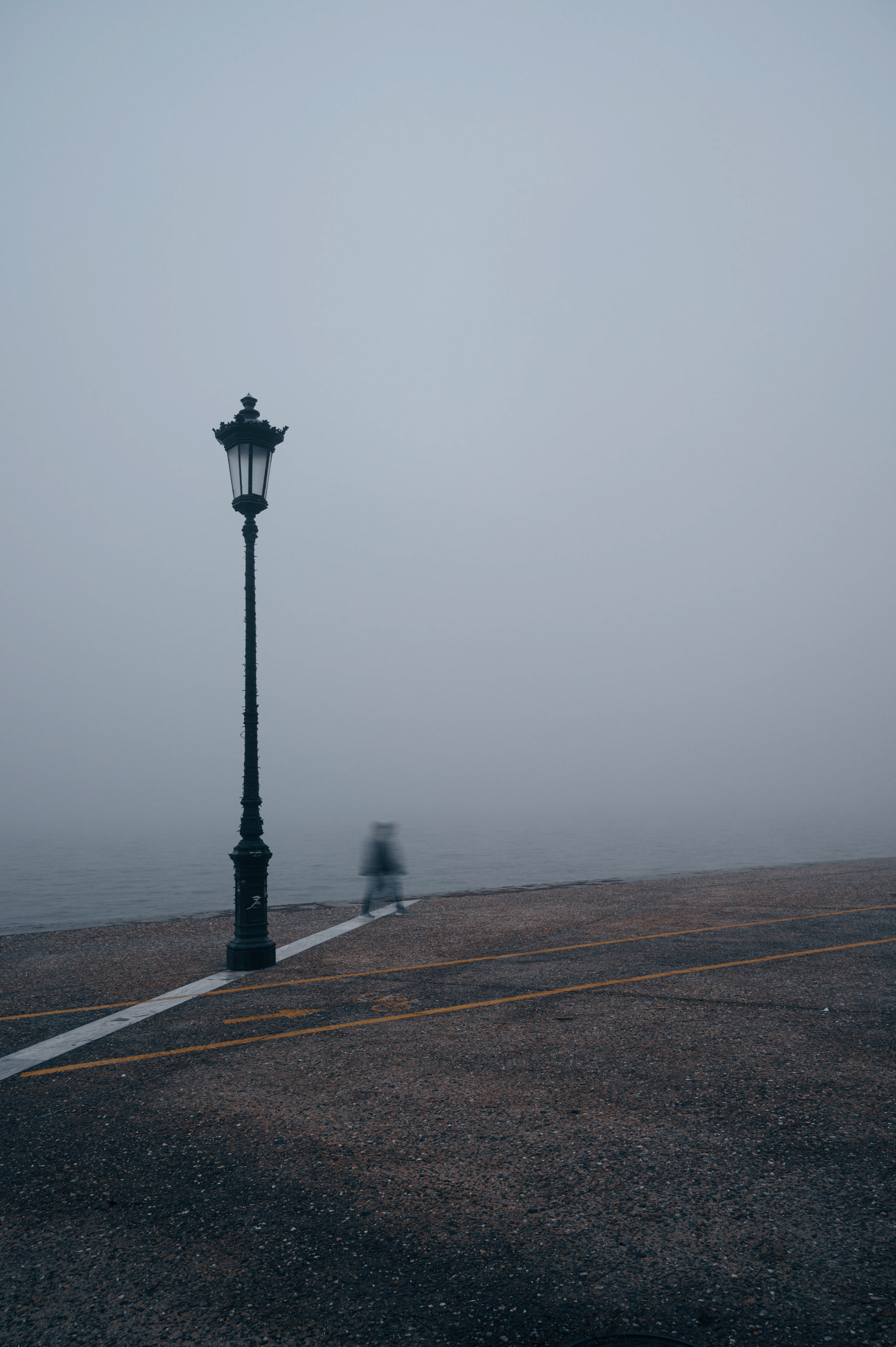 A foggy waterfront scene with a single ornate lamppost standing tall, and a blurred figure walking in the distance..