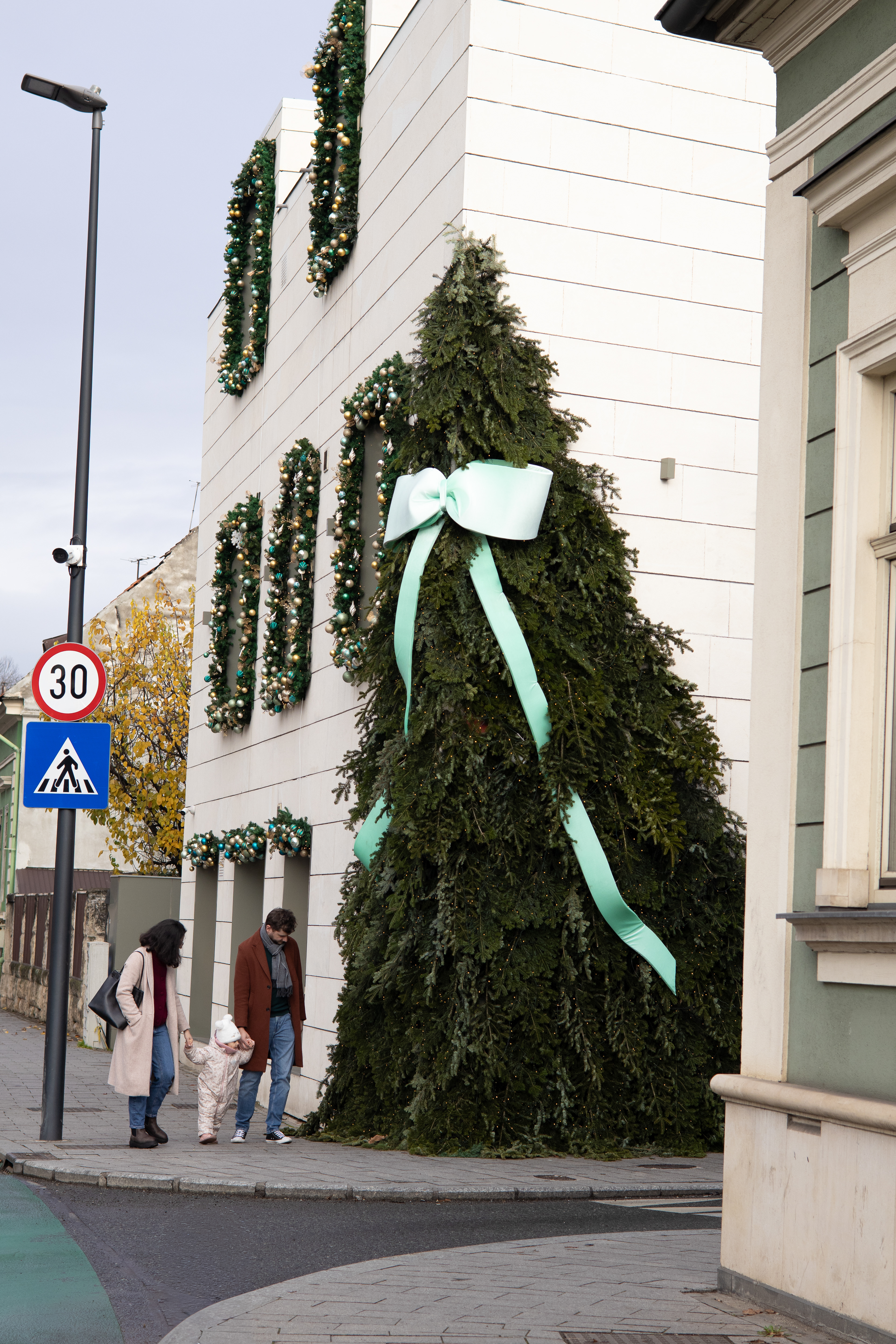 Clădire decorată de sărbători de iarnă, lângă parcul central din Cluj-Napoca. În cadru sunt în trecere o familie cu un copil de mână.