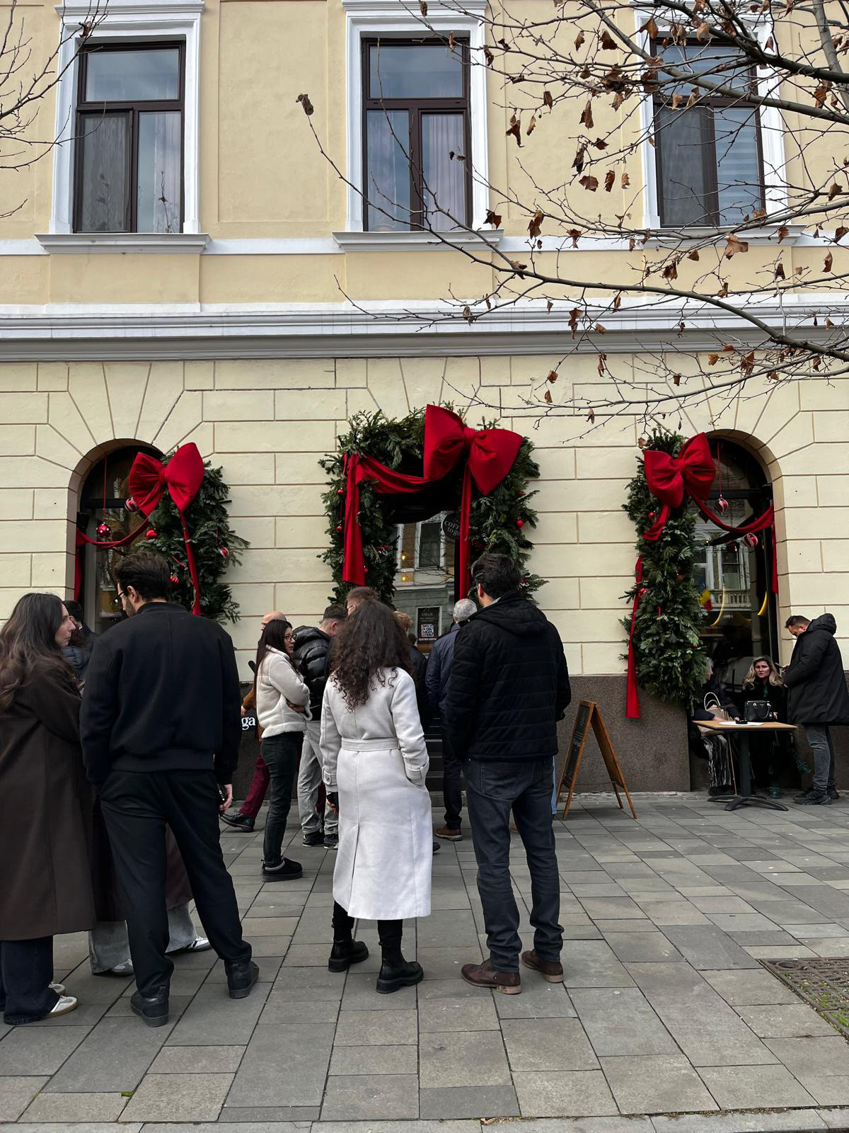 Tineri în fața unei cafenele, stau și discută. În fundal, ferestrele sunt decorate de Crăciun cu fundițe roșii.