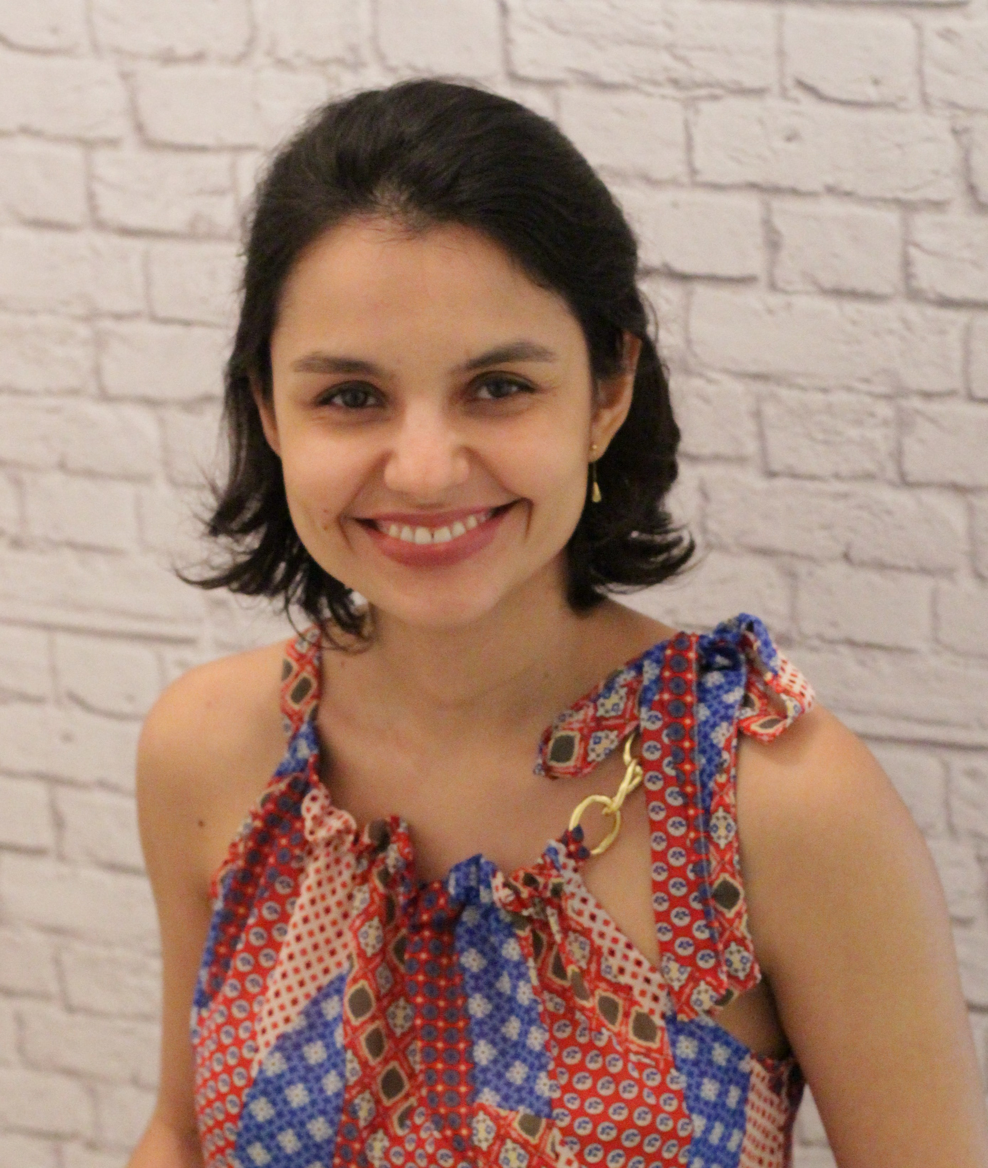 Fotografia de uma mulher branca, de cabelos escuros, acima dos ombros. Ela usa uma blusinha colorida, sem mangas, e está sorrindo.