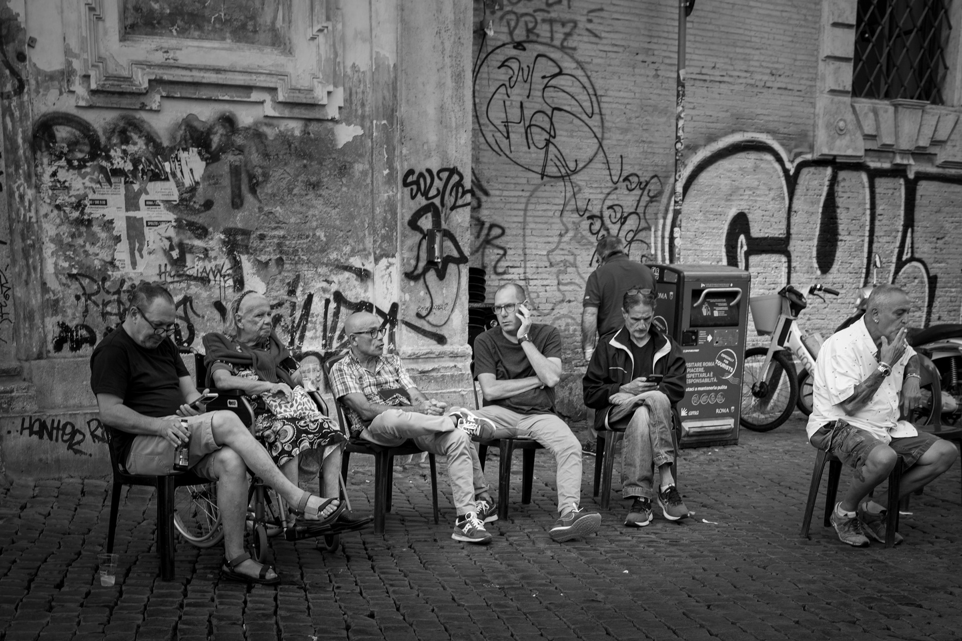 In seduta – un gruppo di uomini seduti in fila davanti a un muro di graffiti a Trastevere, Roma. Fotografia in bianco e nero di Fabio Alimonti.