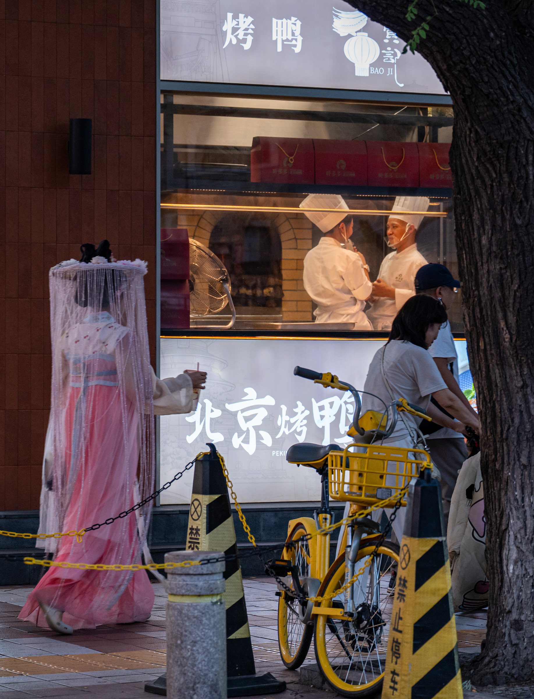 Pechino: una ragazza in hanfu con il velo passa davanti alla vetrina di un ristorante di anatra laccata, accanto a una bicicletta gialla