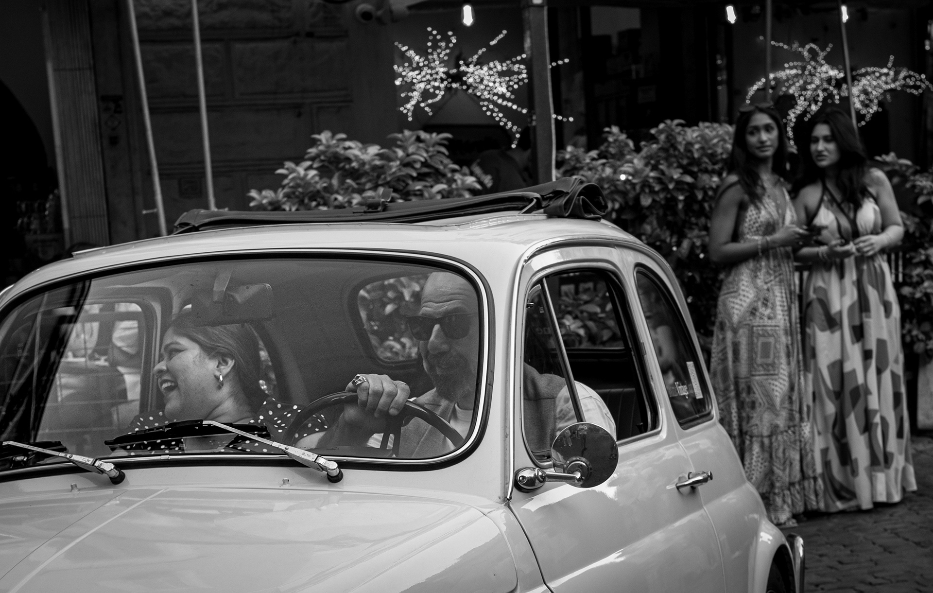 500 risate – bianco e nero: in una vecchia Fiat 500 una donna ride mentre l'uomo alla guida sogghigna con l'anello al mignolo; dietro, due ragazze in abito lungo li guardano passare a Trastevere. Fotografia di Fabio Alimonti.