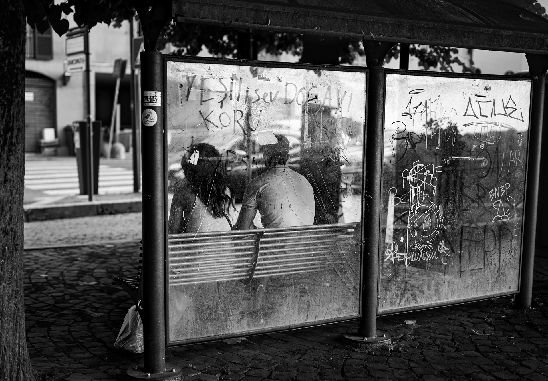 Due persone sedute alla fermata dell'autobus di Nemi, viste attraverso il vetro graffiato e coperto di scritte.