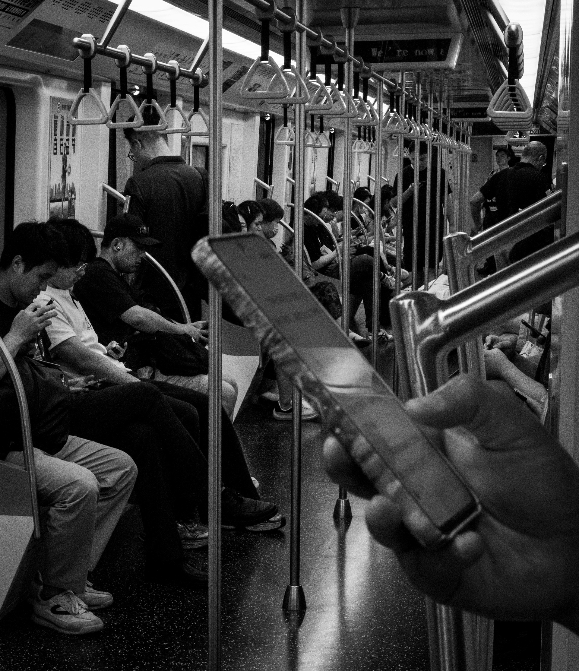 Vagone della metropolitana di Shanghai: passeggeri seduti e in piedi, ognuno chino sul proprio telefono; in primo piano una mano stringe uno smartphone — Fabio Alimonti
